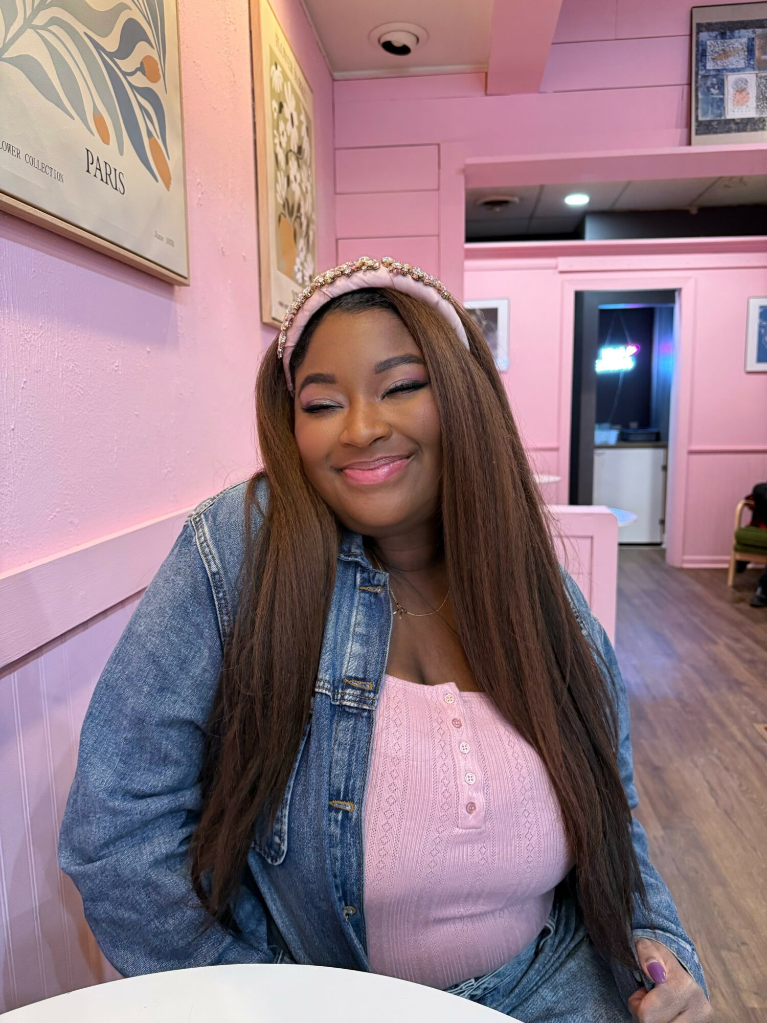 Young woman with long hair smiling, wearing a pink headband, pink top, and denim jacket, sitting in a pink-themed room.