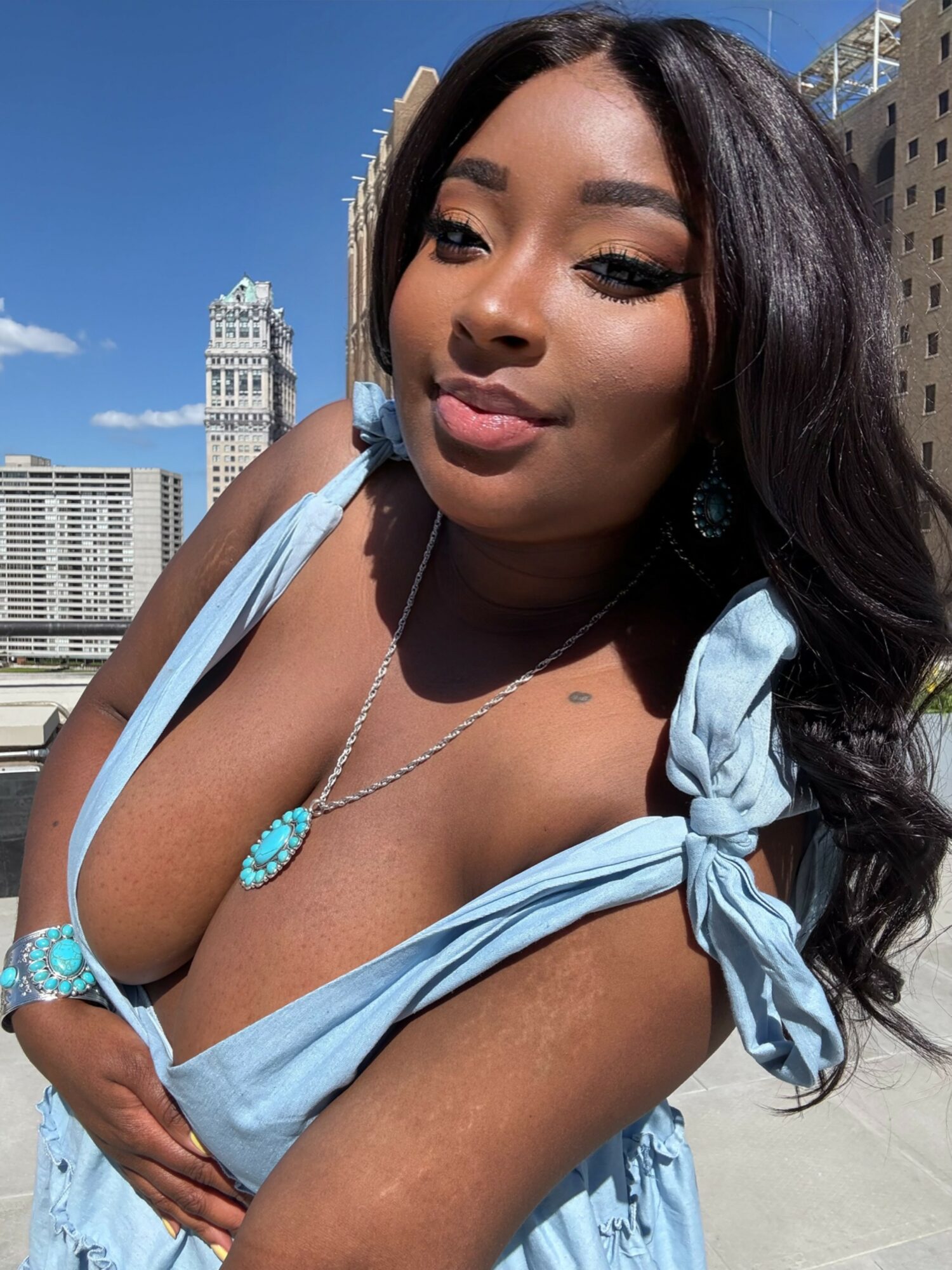Young woman with long dark hair wearing a light blue dress and jewelry, outdoors with city buildings in background.
