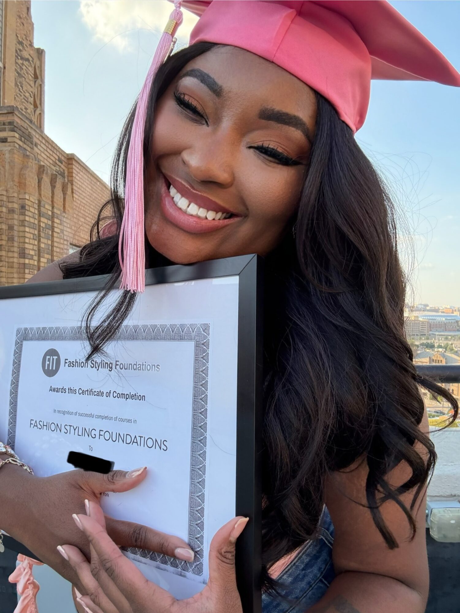 Young woman in pink graduation cap smiling, holding a certificate, outdoors with a building and sky in background.