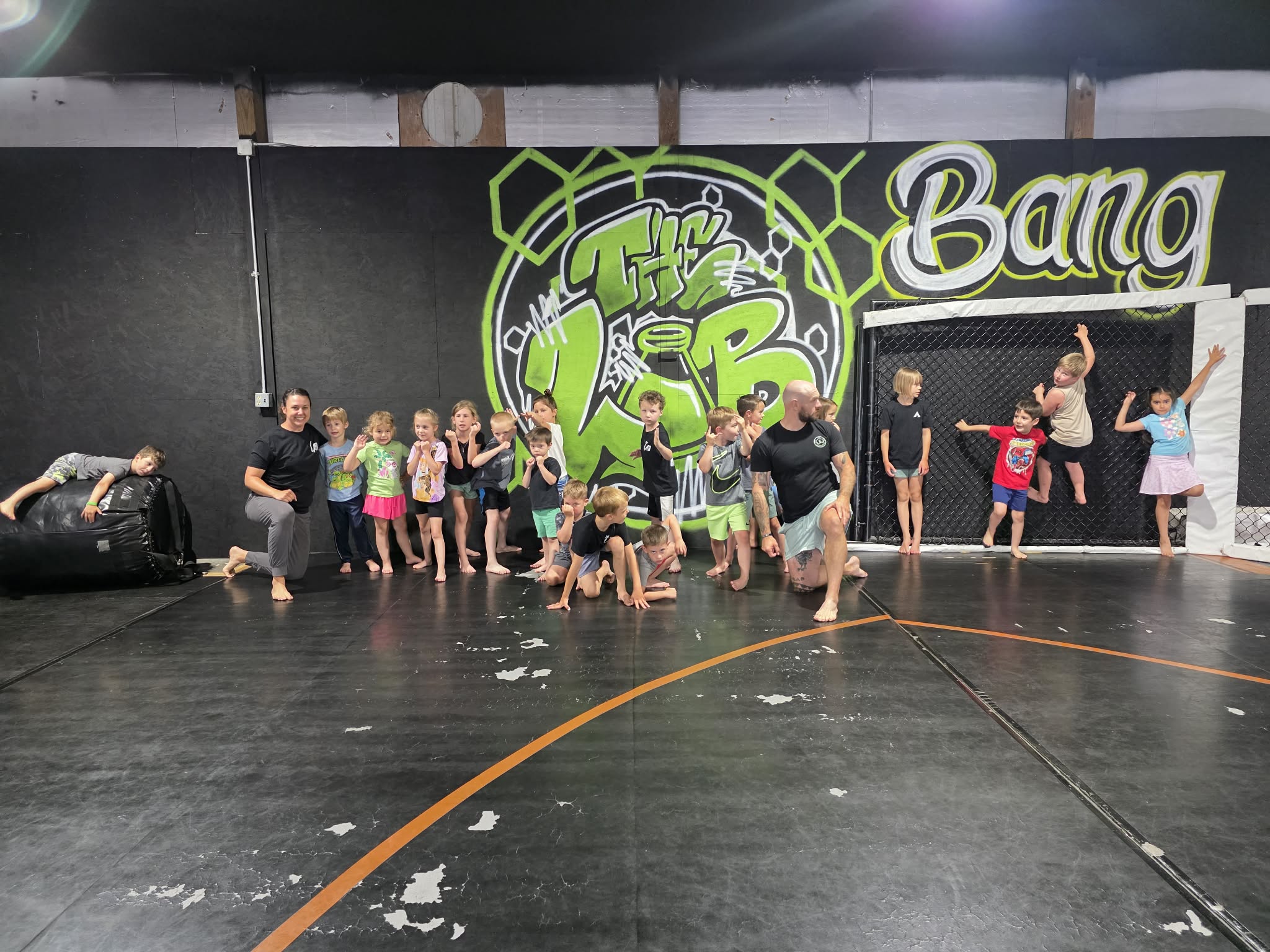 Group of children and adults in a gym with graffiti-style wall art, some children are jumping or sitting on the floor.