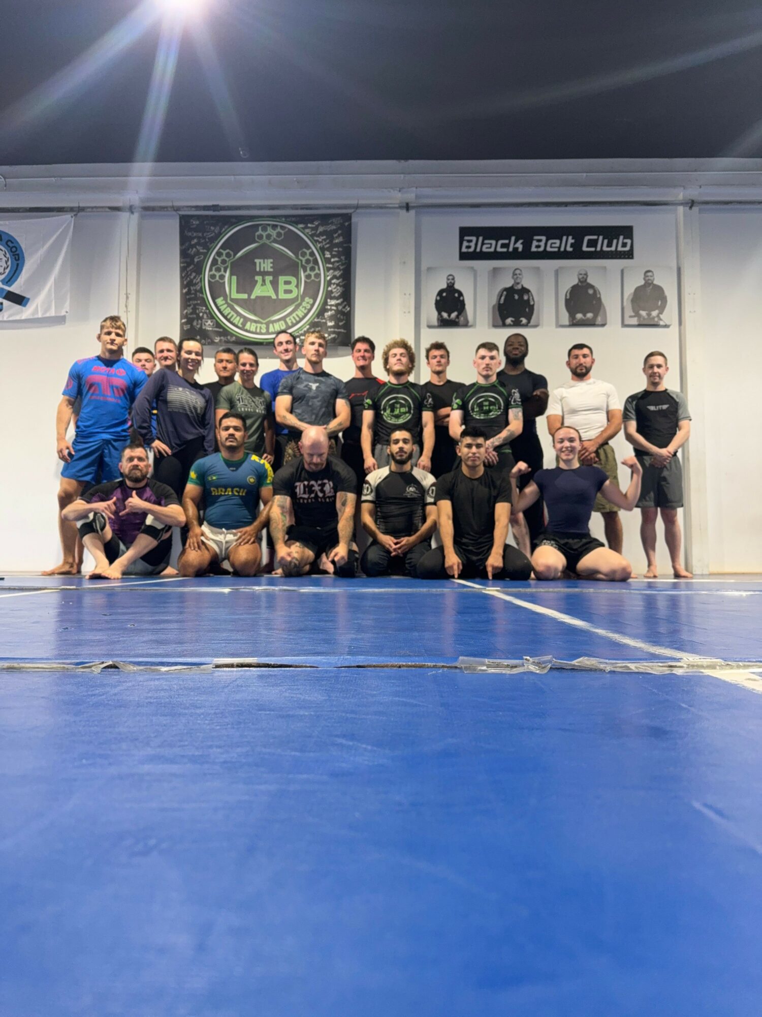 Group of people in a gym, some sitting and some standing, with a blue floor and banners on the wall.