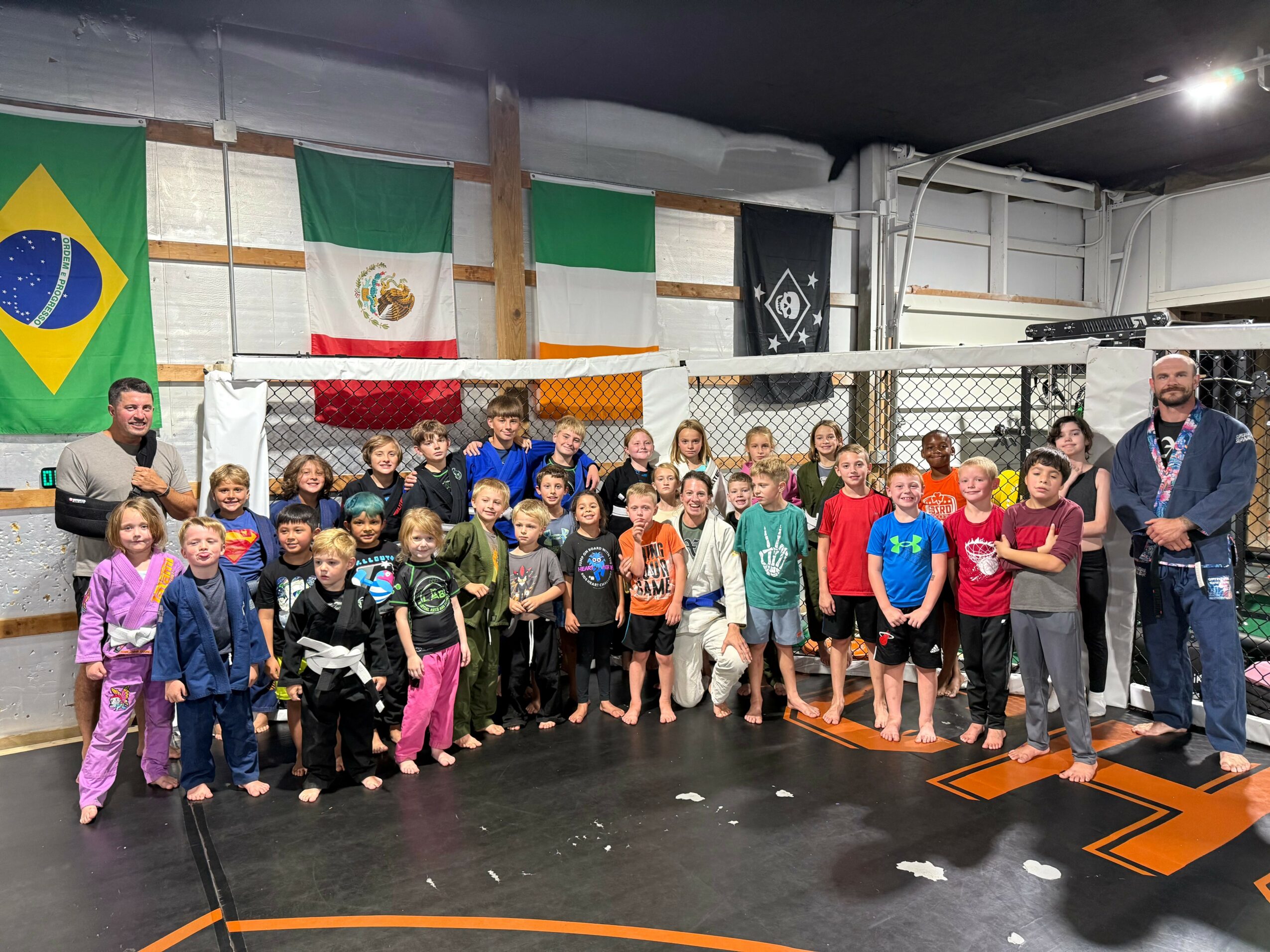 Group of children and adults in a gym with flags of Brazil, Mexico, and others hanging on the wall.