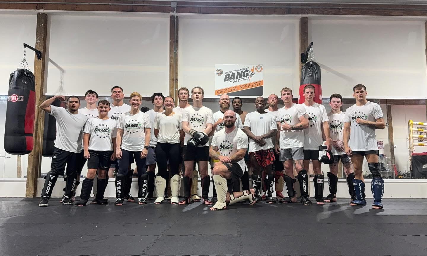 Group of people in boxing gym, some with boxing gloves, standing in front of punching bags, smiling, with a banner in the background.