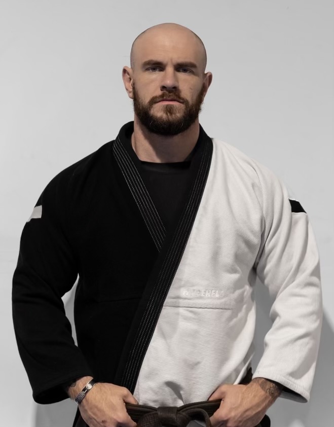 Bald man with a beard wearing a black and white martial arts gi, standing with hands on hips against a plain background.