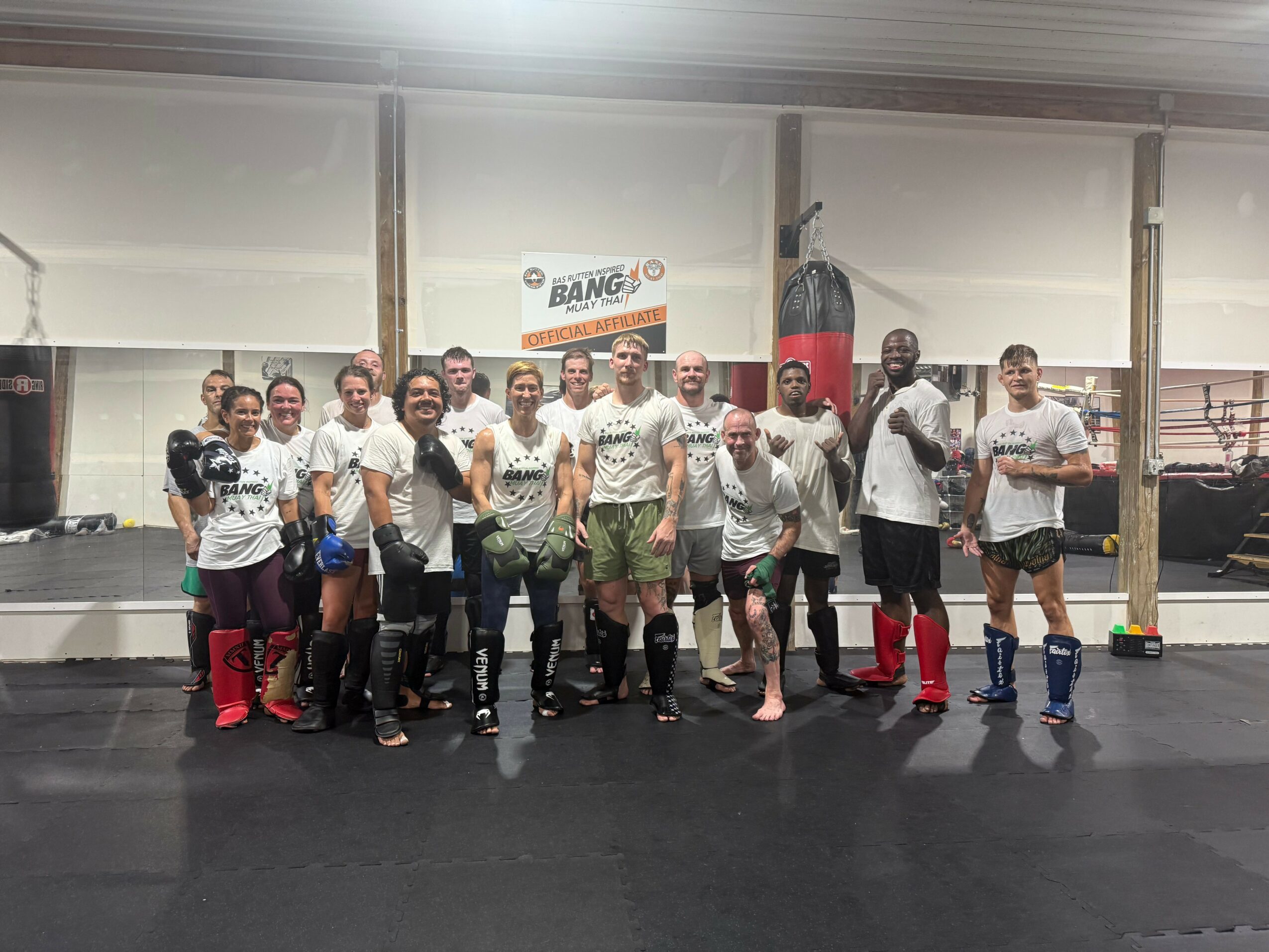 Group of people in boxing gym, some wearing boxing gloves and shorts, standing in front of punching bag, smiling.