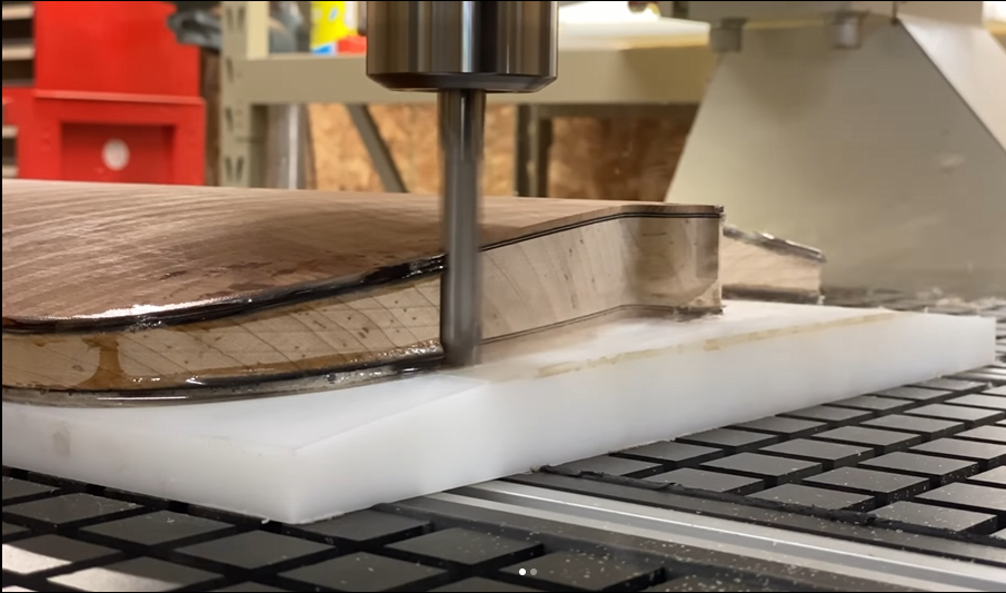 A CNC machine cuts a wooden guitar body on a white surface with a grid pattern.