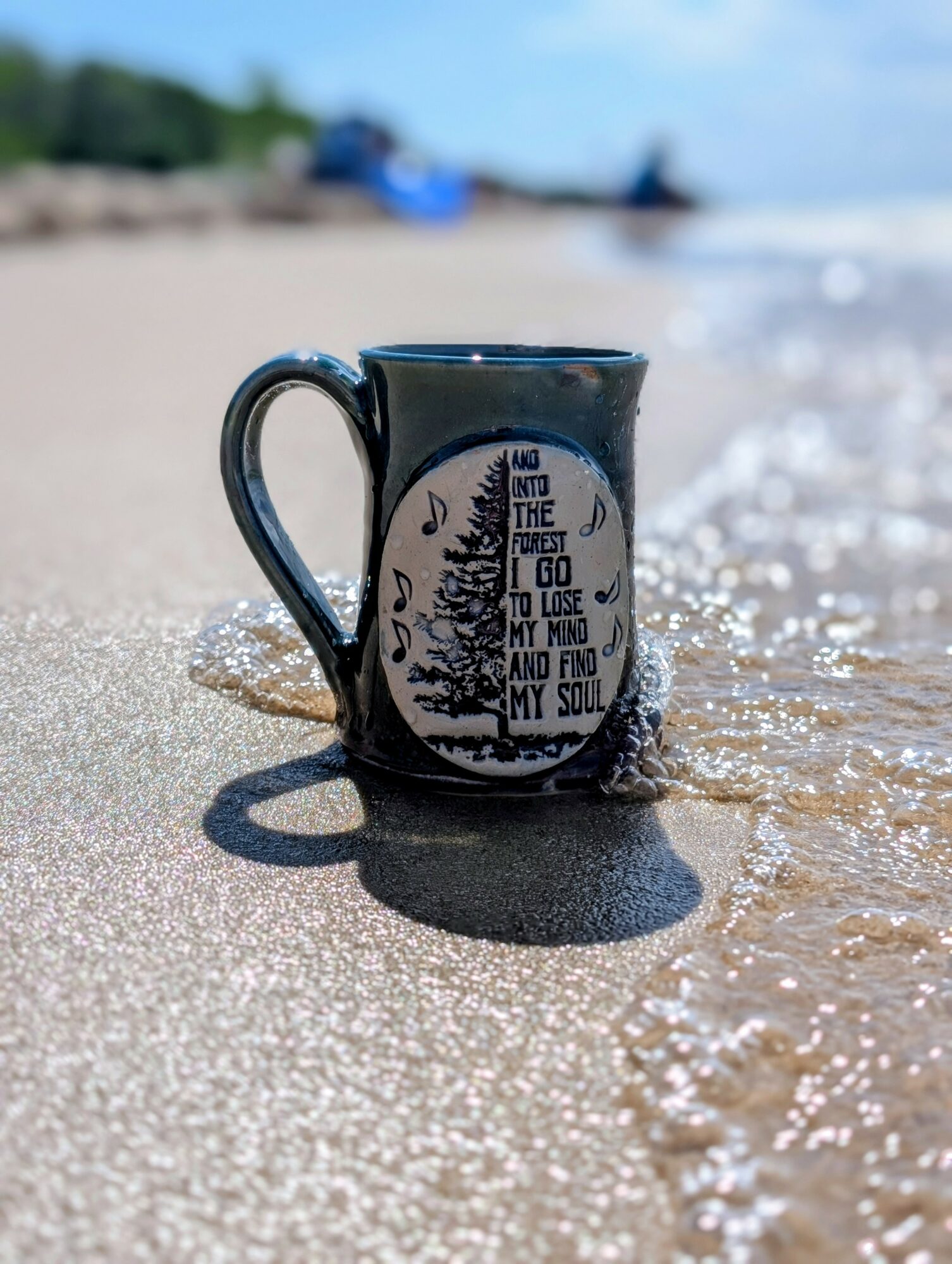 A black mug with a quote and design, placed on wet sand at the beach, with water and sky in background.
