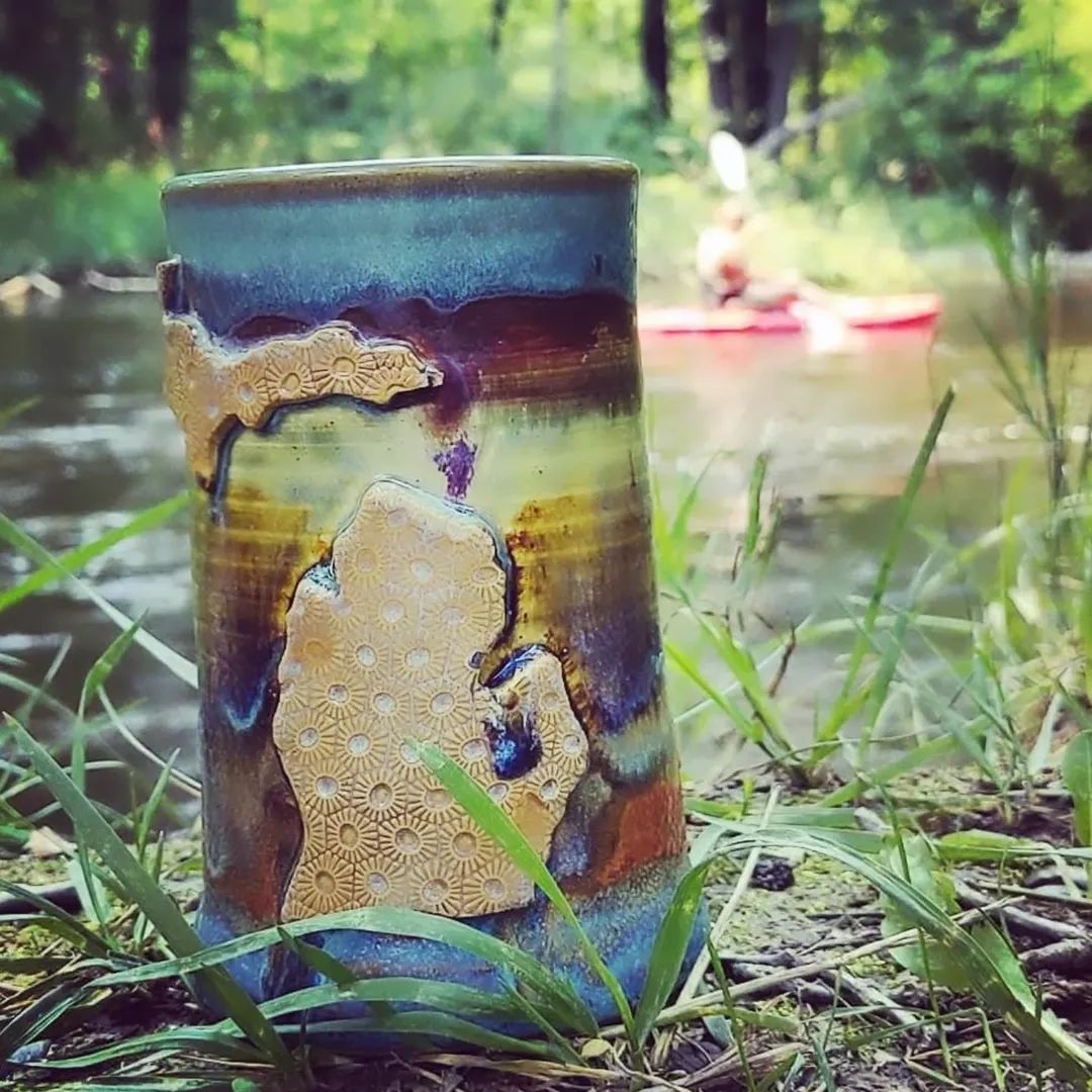 A colorful ceramic mug on grass near a river with a person kayaking in the background.