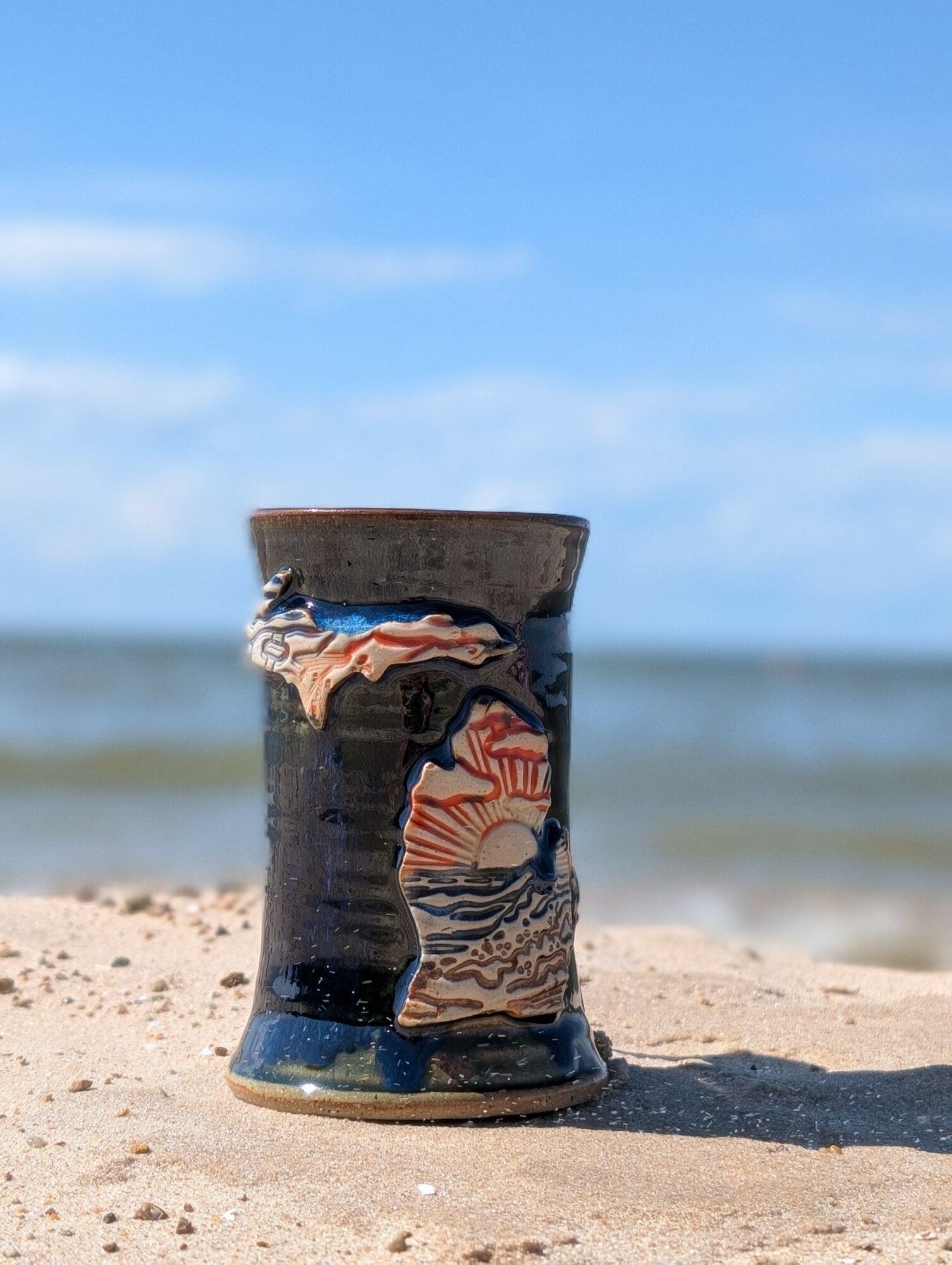 Ceramic cup on sandy beach with ocean and blue sky in background.