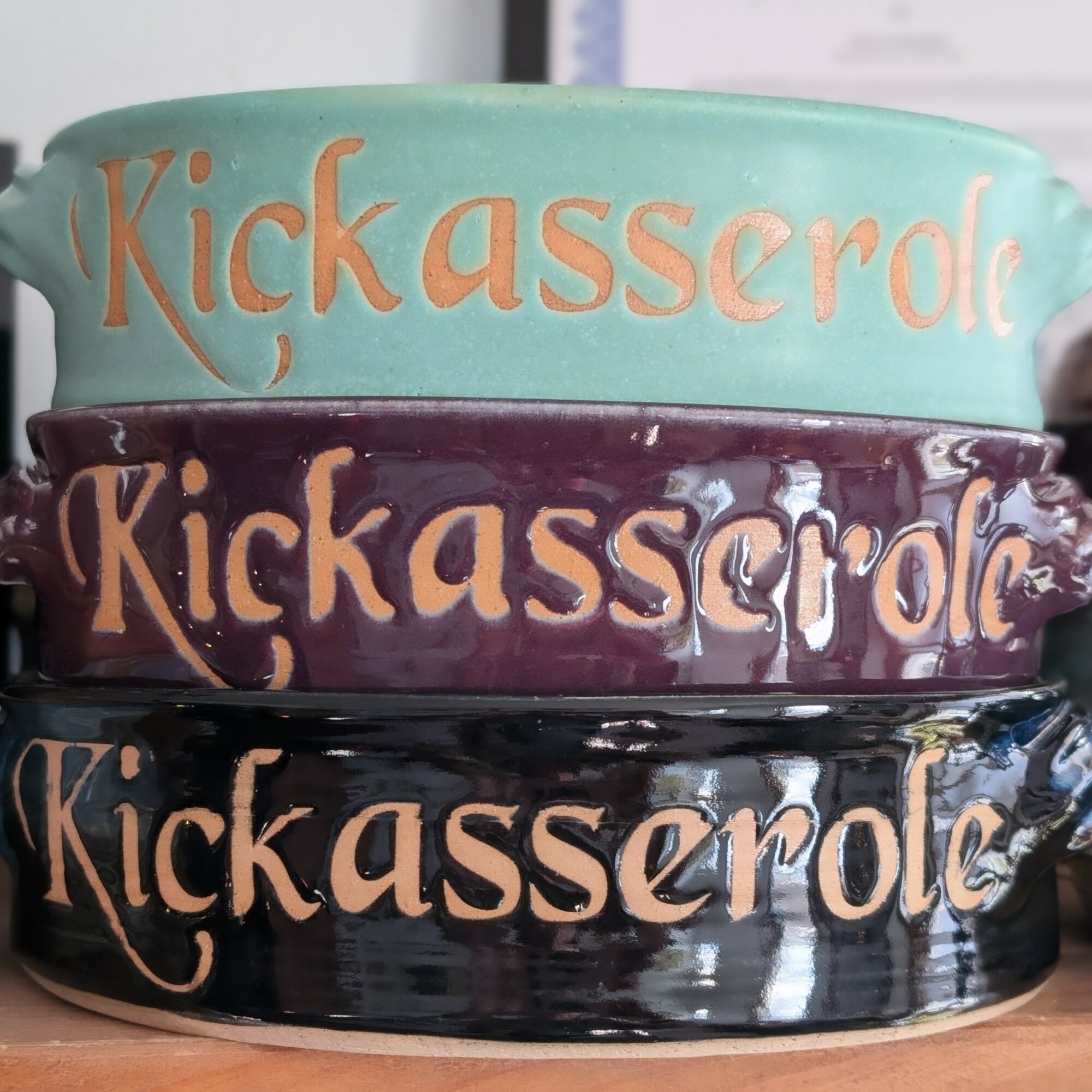 Three stacked ceramic bowls with the word 'Kickassercle' written on each in different colors, from top to bottom: light blue, purple, black.