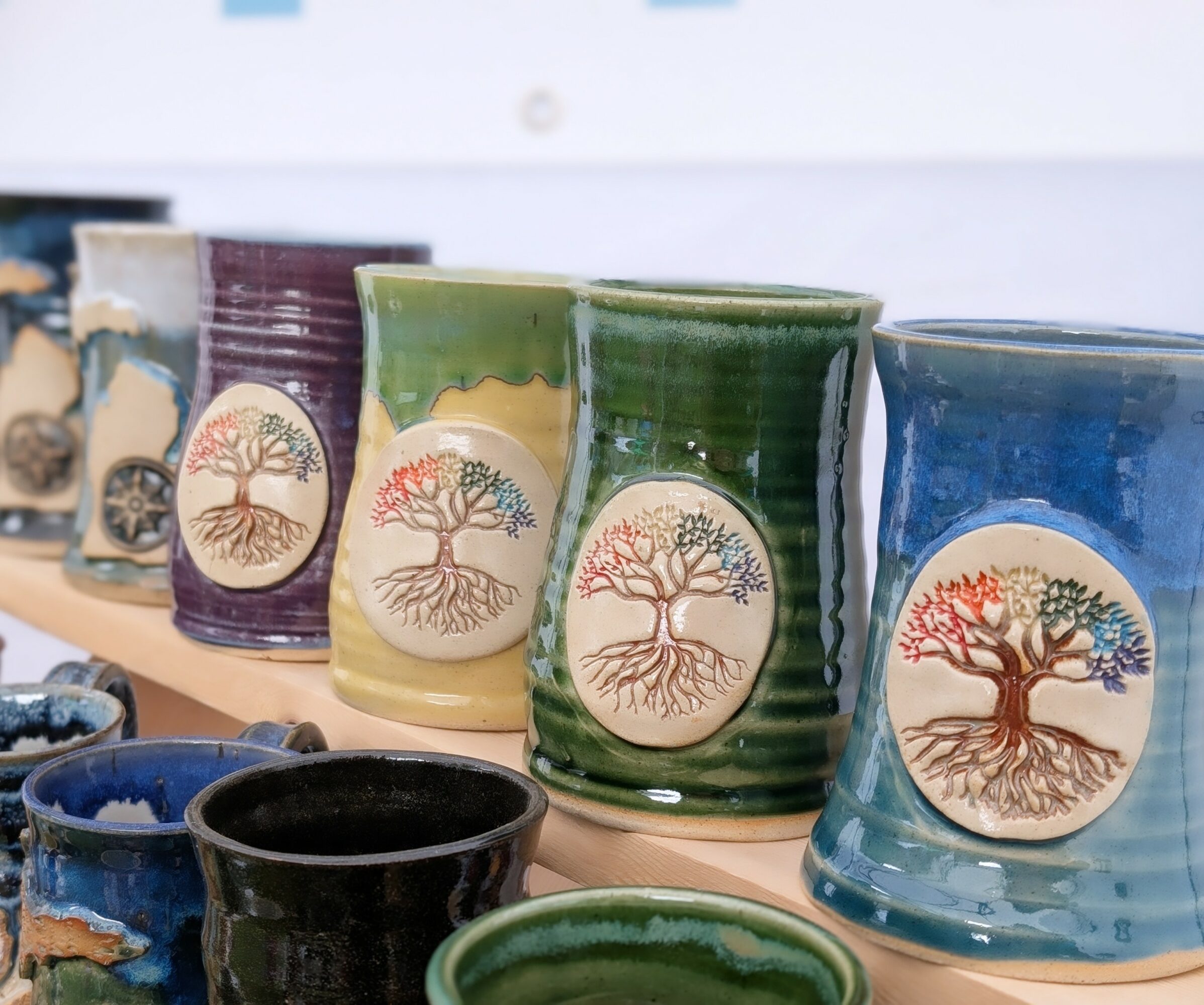 Colorful ceramic cups with tree designs, arranged on a shelf, in various colors including purple, yellow, green, and blue.