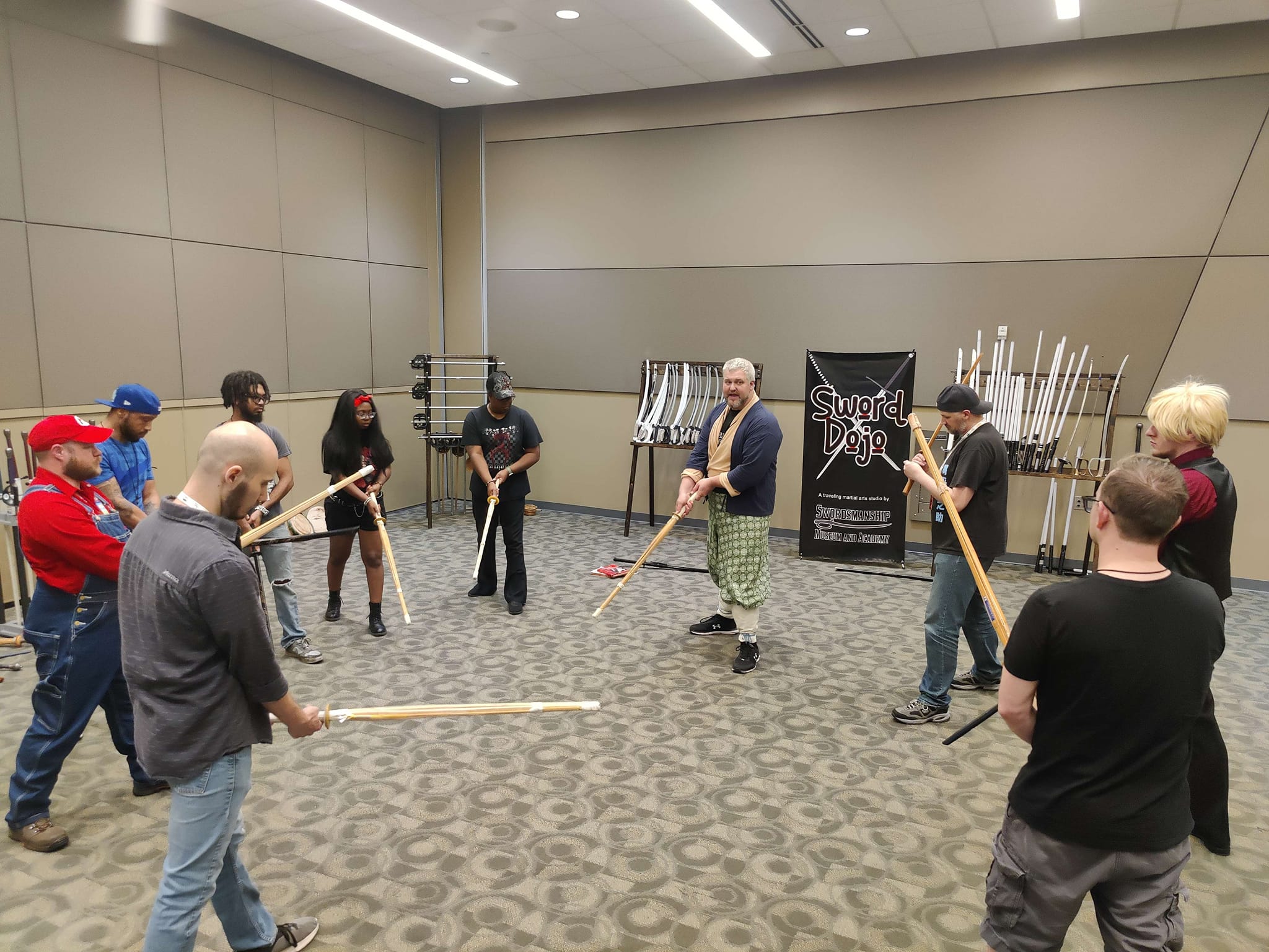 Group of people standing in a circle holding long wooden sticks in a room with gray walls and carpeted floor.