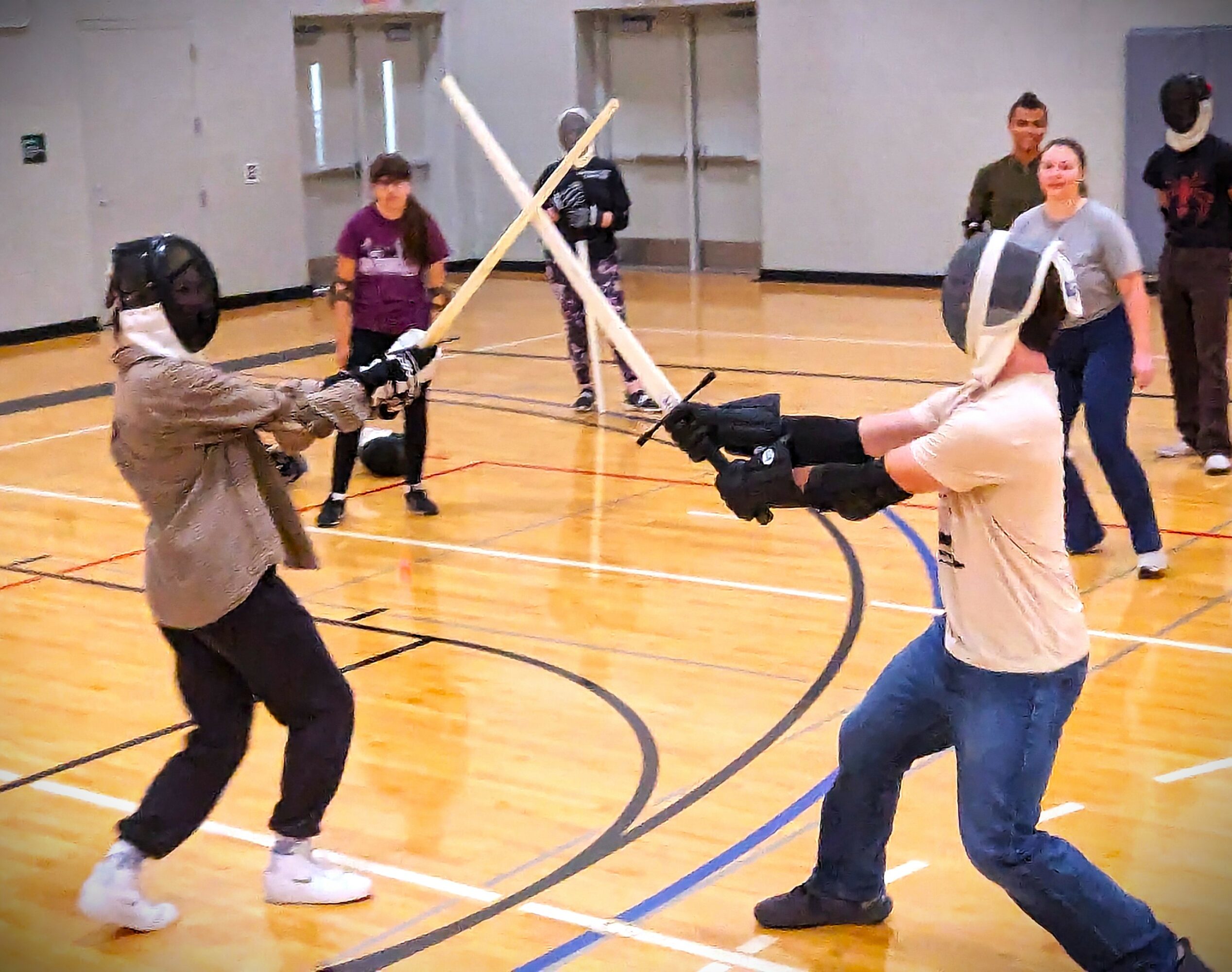 Two people in fencing gear sparring with swords in a gymnasium, others watch in the background.