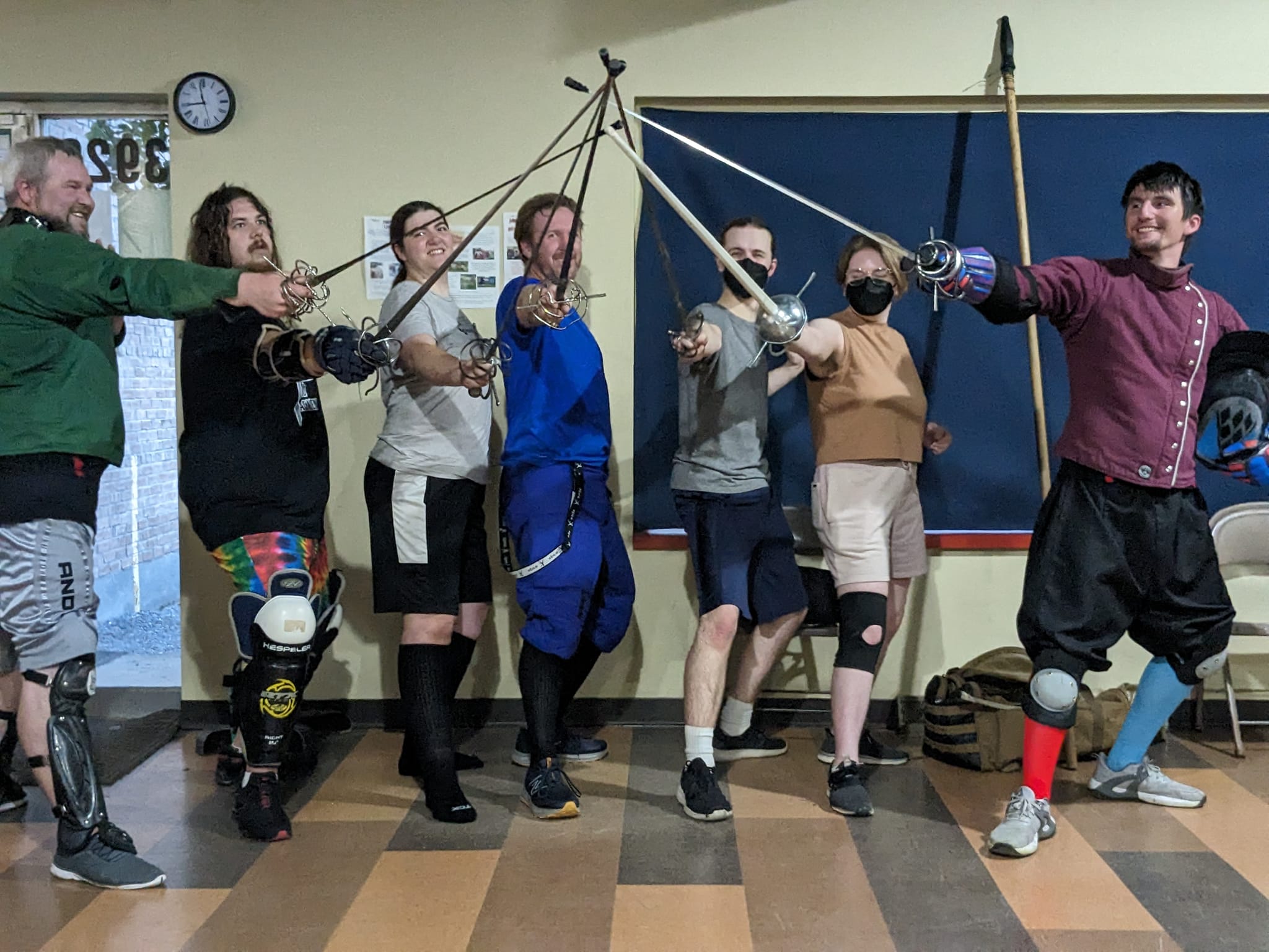 Group of people practicing fencing indoors, holding swords and wearing casual clothing, with a blackboard and clock in the background.