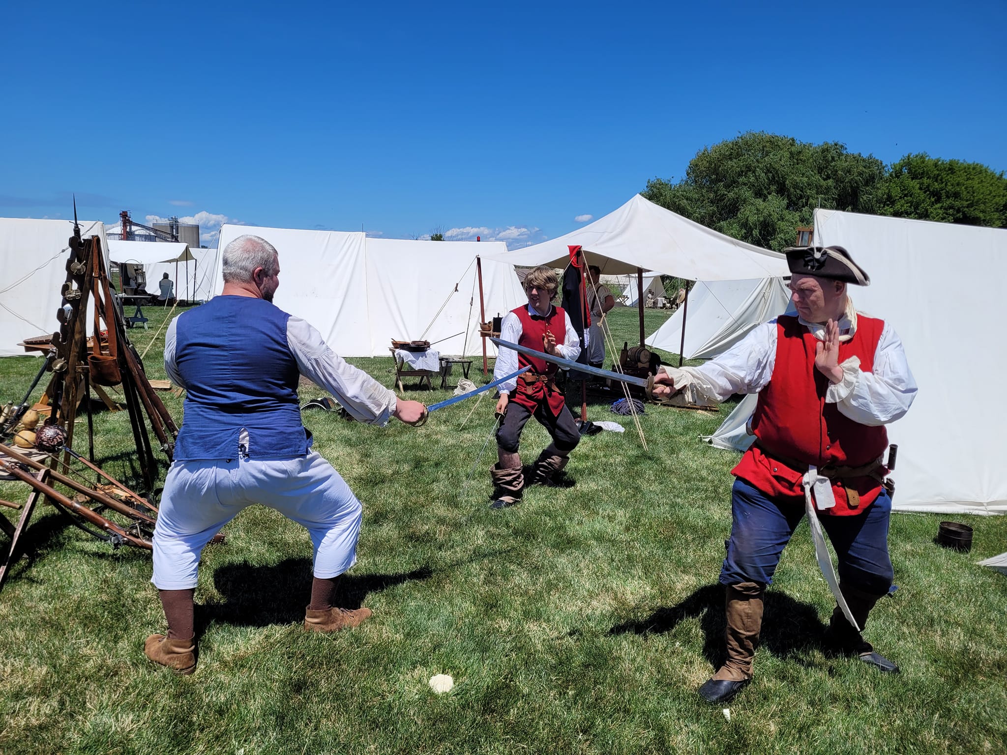 Three people in pirate costumes practicing sword fighting outdoors with tents and trees in the background.