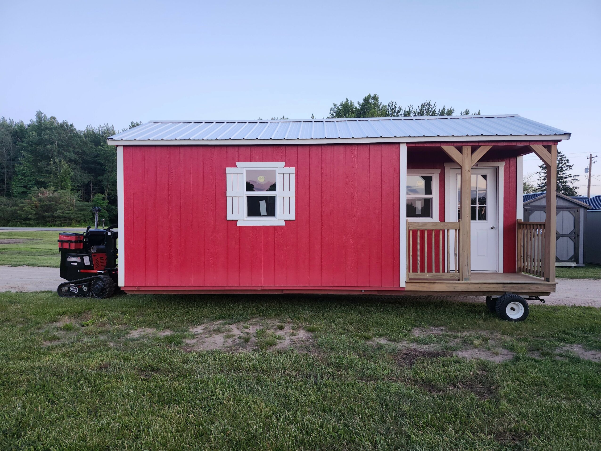 Red tiny house on wheels with white window shutters and a porch, set on grass with trees in background.