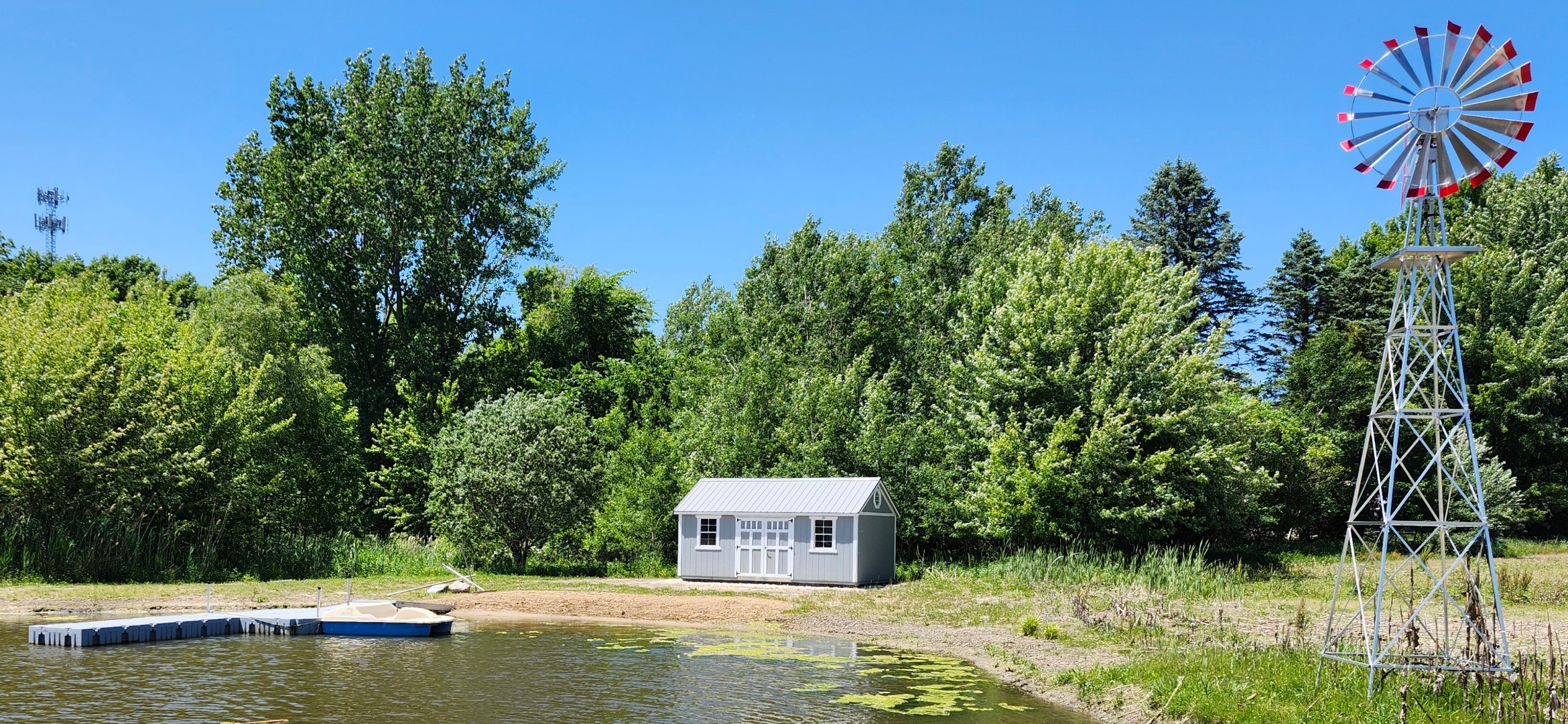 A small white building near water, surrounded by green trees and a blue sky, with a windmill on the right.