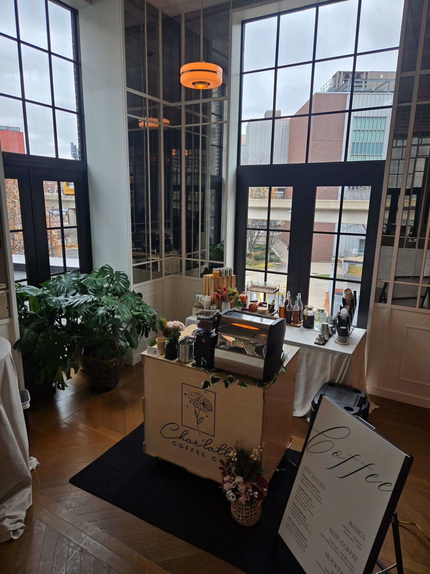 Interior view of a room with large windows, a table with plants, books, and a menu, and a cityscape outside.