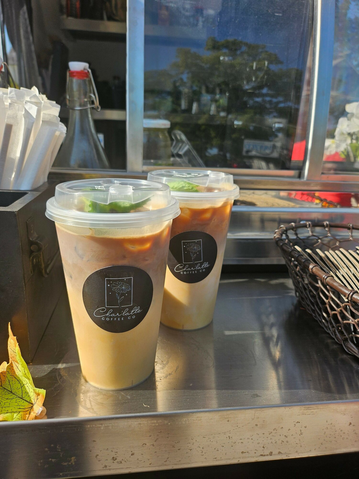 Two iced beverages in plastic cups with black labels, placed on a metal counter near a window, with a plastic container and a basket nearby.