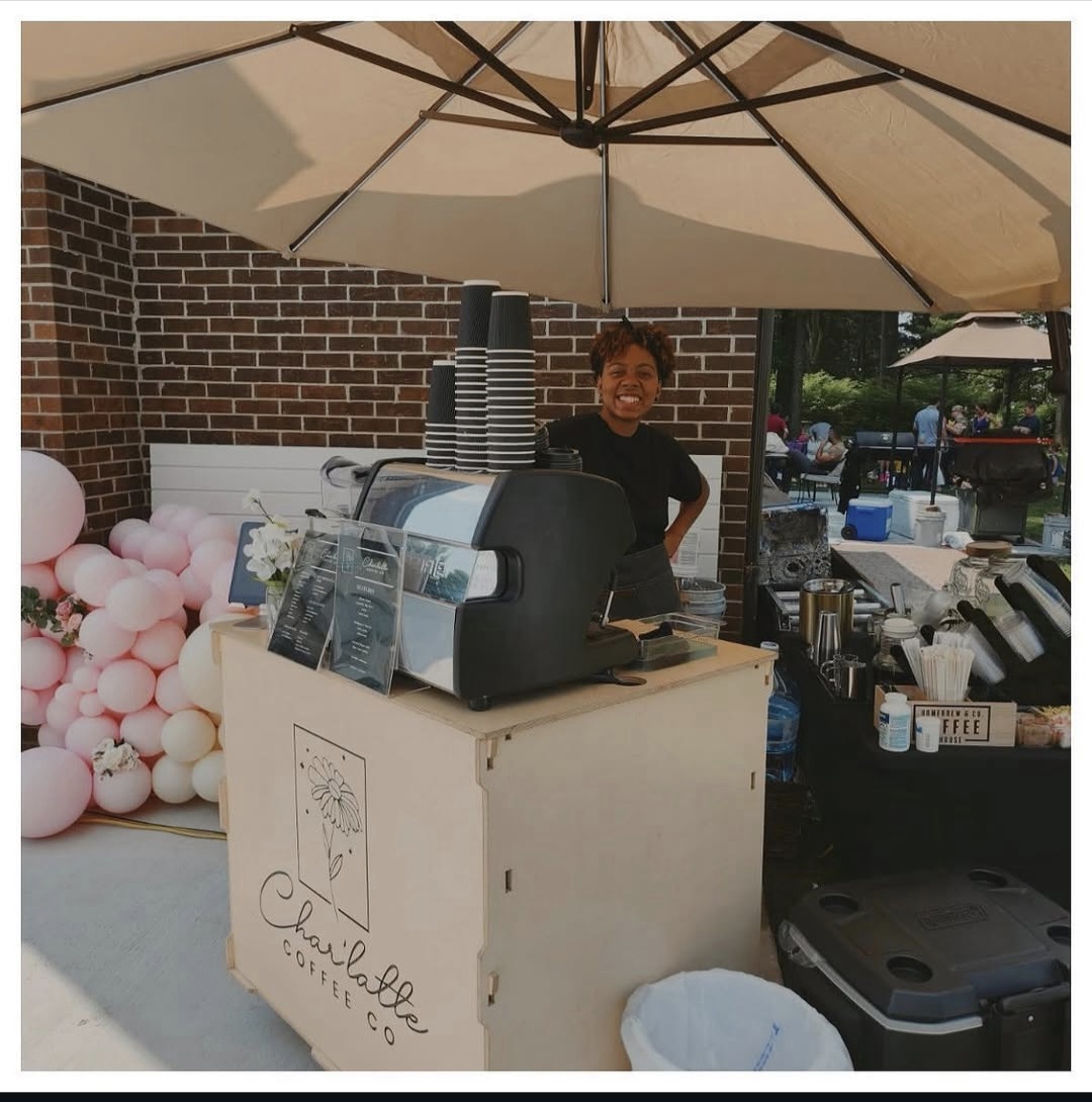 Person standing behind a coffee stand under a large umbrella, with balloons and outdoor setting in background.