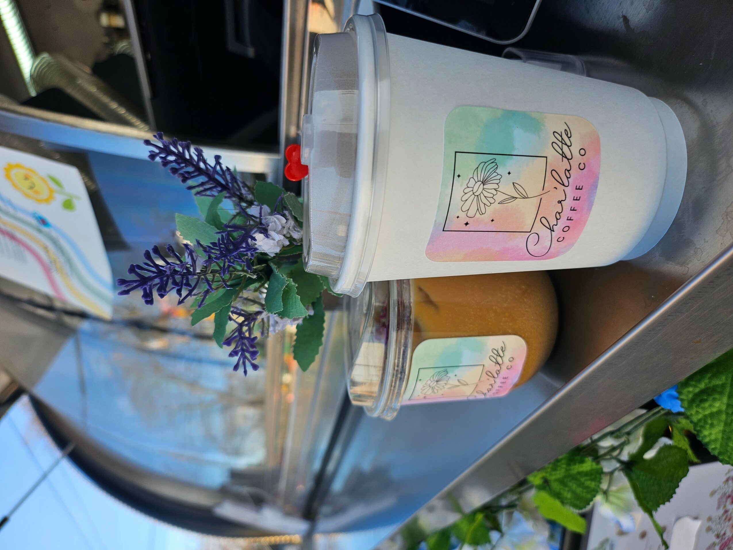 A white cup with a colorful label and a small jar with a similar label, placed on a surface with a plant nearby.