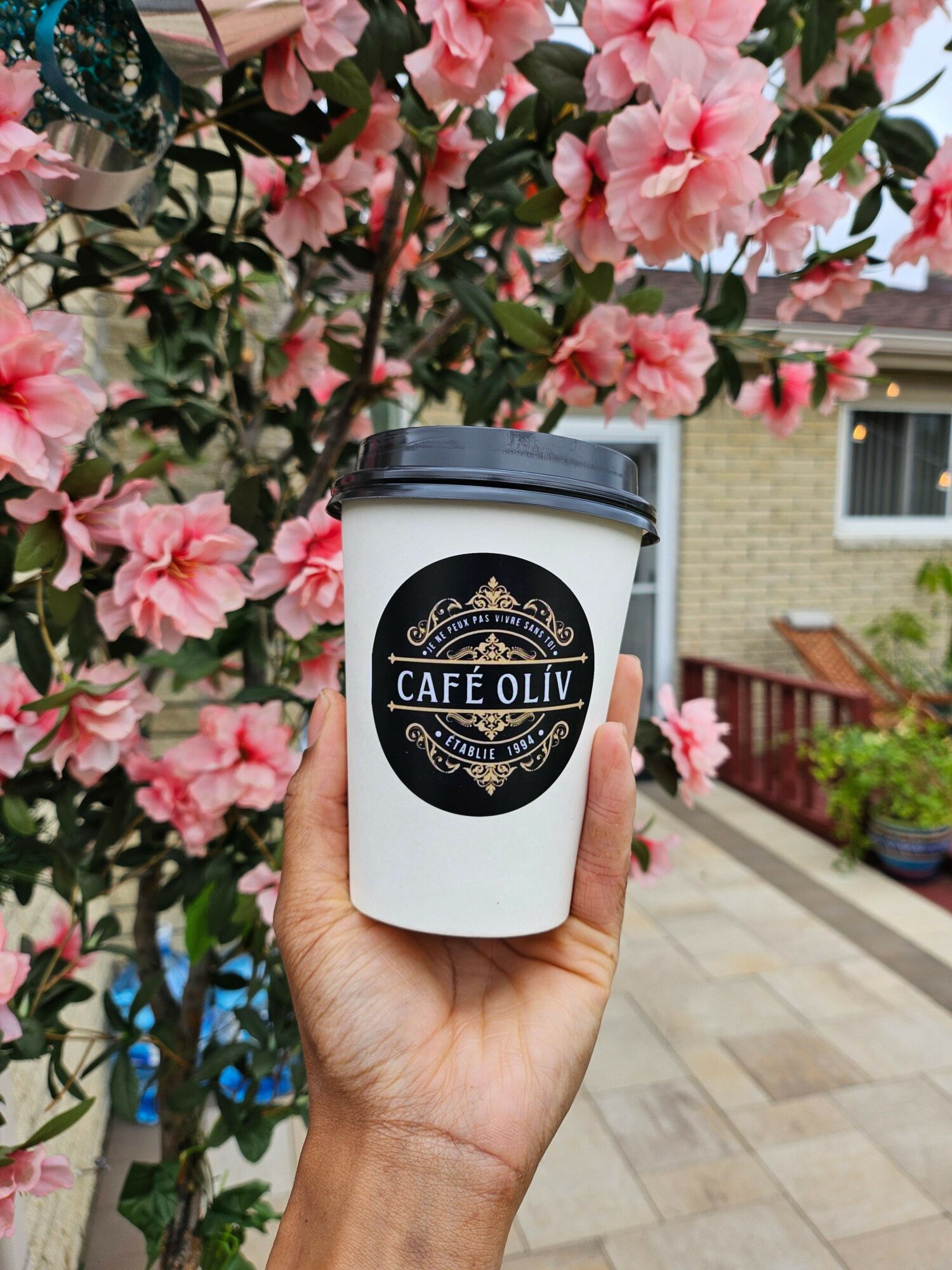 Person holding a coffee cup with a black and white logo, pink flowers in background, outdoor setting