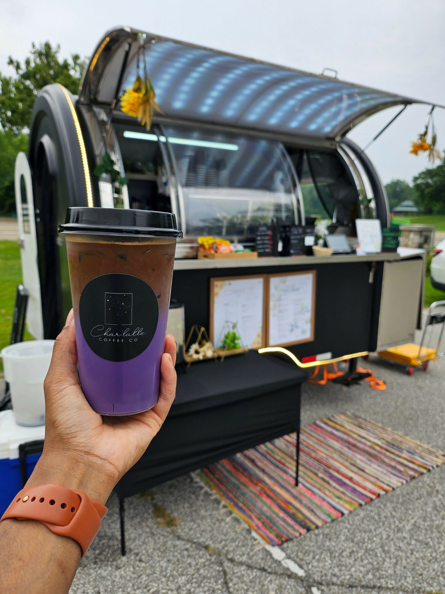 Person holding a purple cup with a black label, outdoor setup with a black canopy, colorful rug, and display boards.