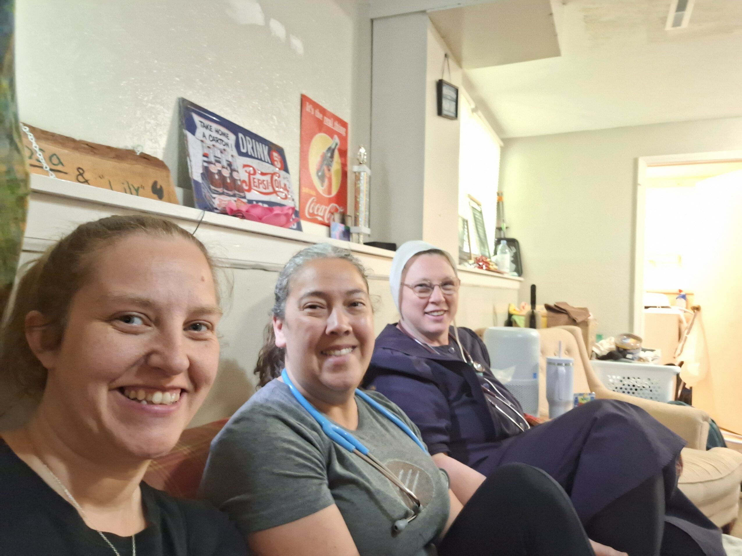 Three women sitting on a couch in a room with shelves and a window, smiling at the camera.