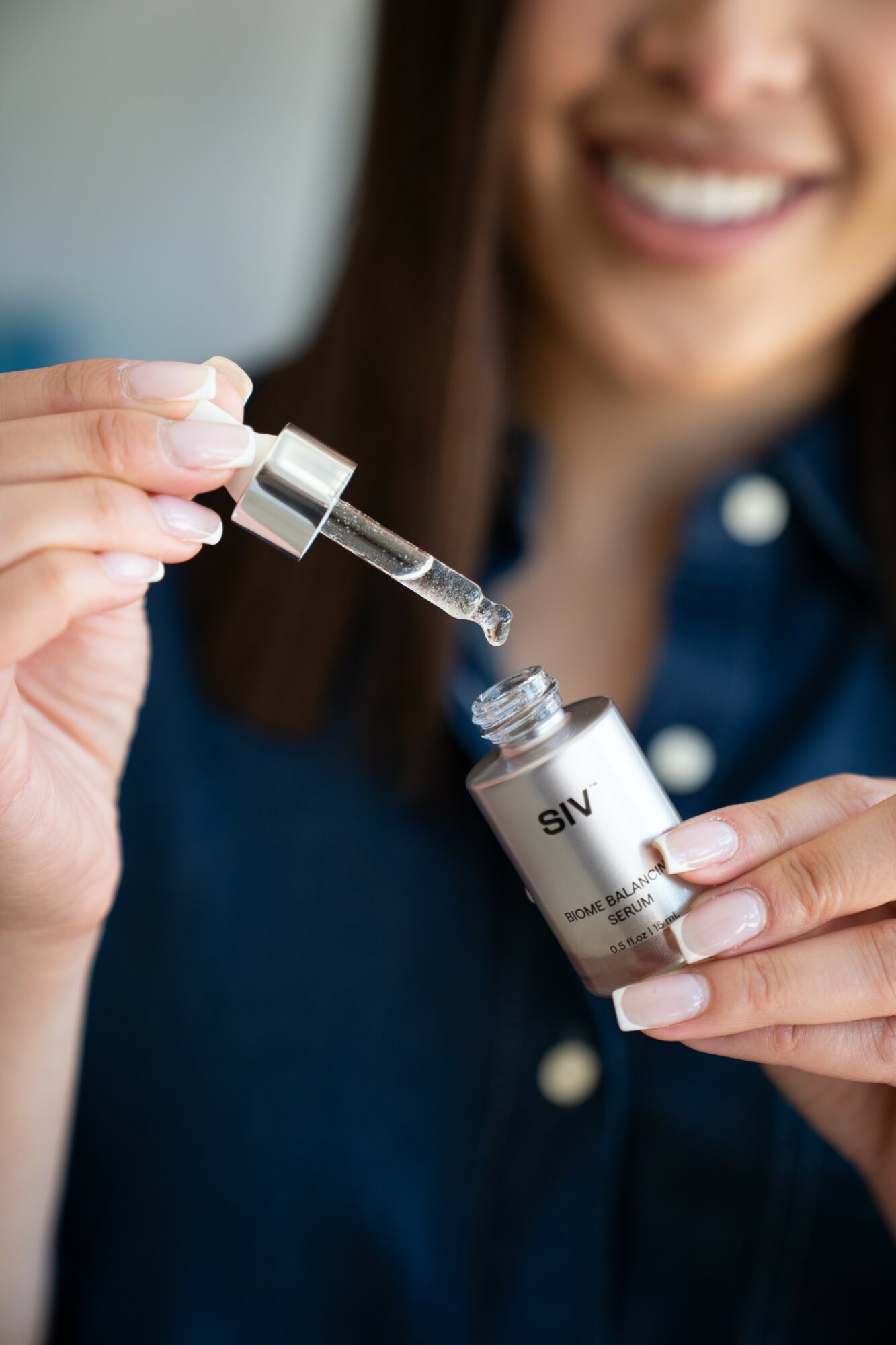 Person holding a dropper and a small bottle, smiling, with focus on hands and dropper, background blurred.