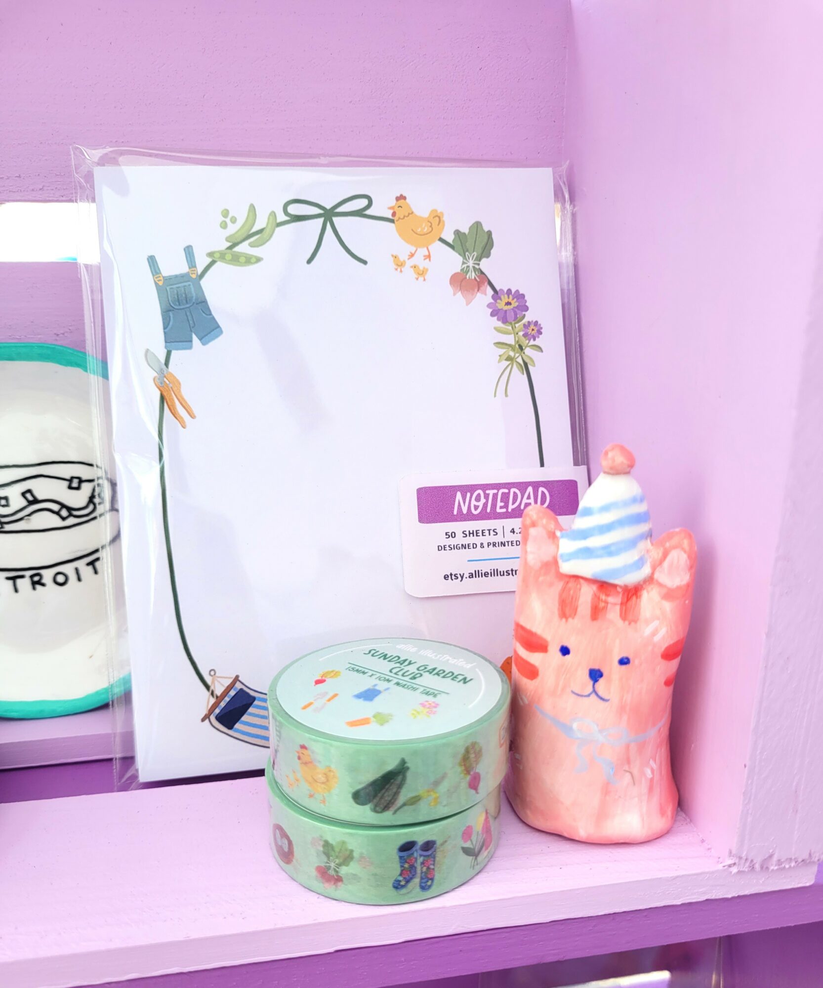 Pink plush cat with a blue and white striped hat, next to a round pastel container, on a pink shelf.