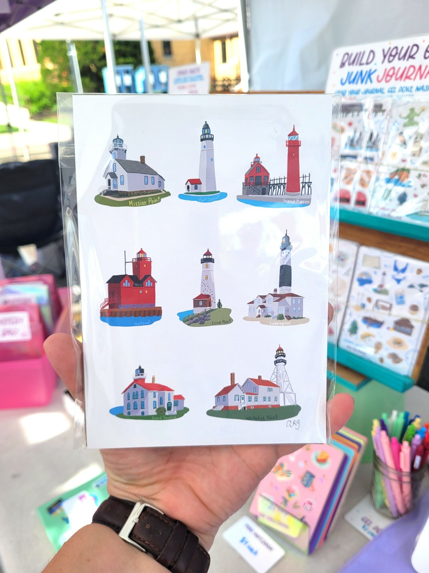 Hand holding a card with illustrations of eight different lighthouses, each with distinct designs and colors.