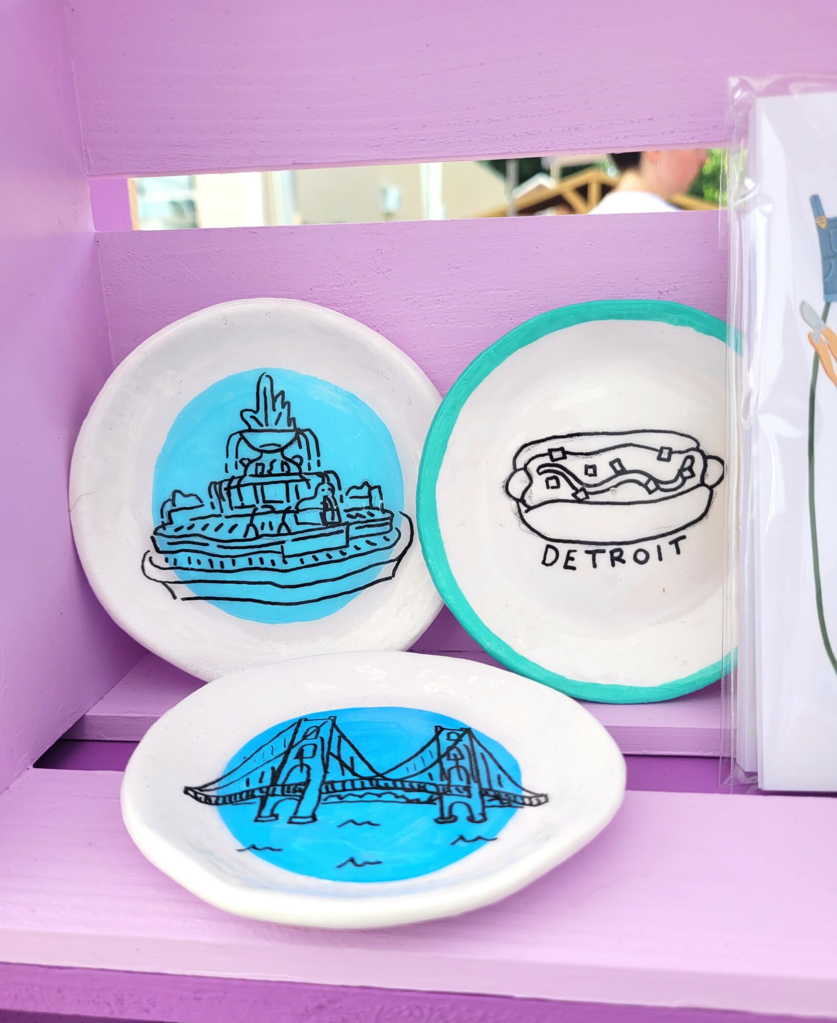 Three small plates with blue and black line drawings of Detroit landmarks, placed on a purple shelf.