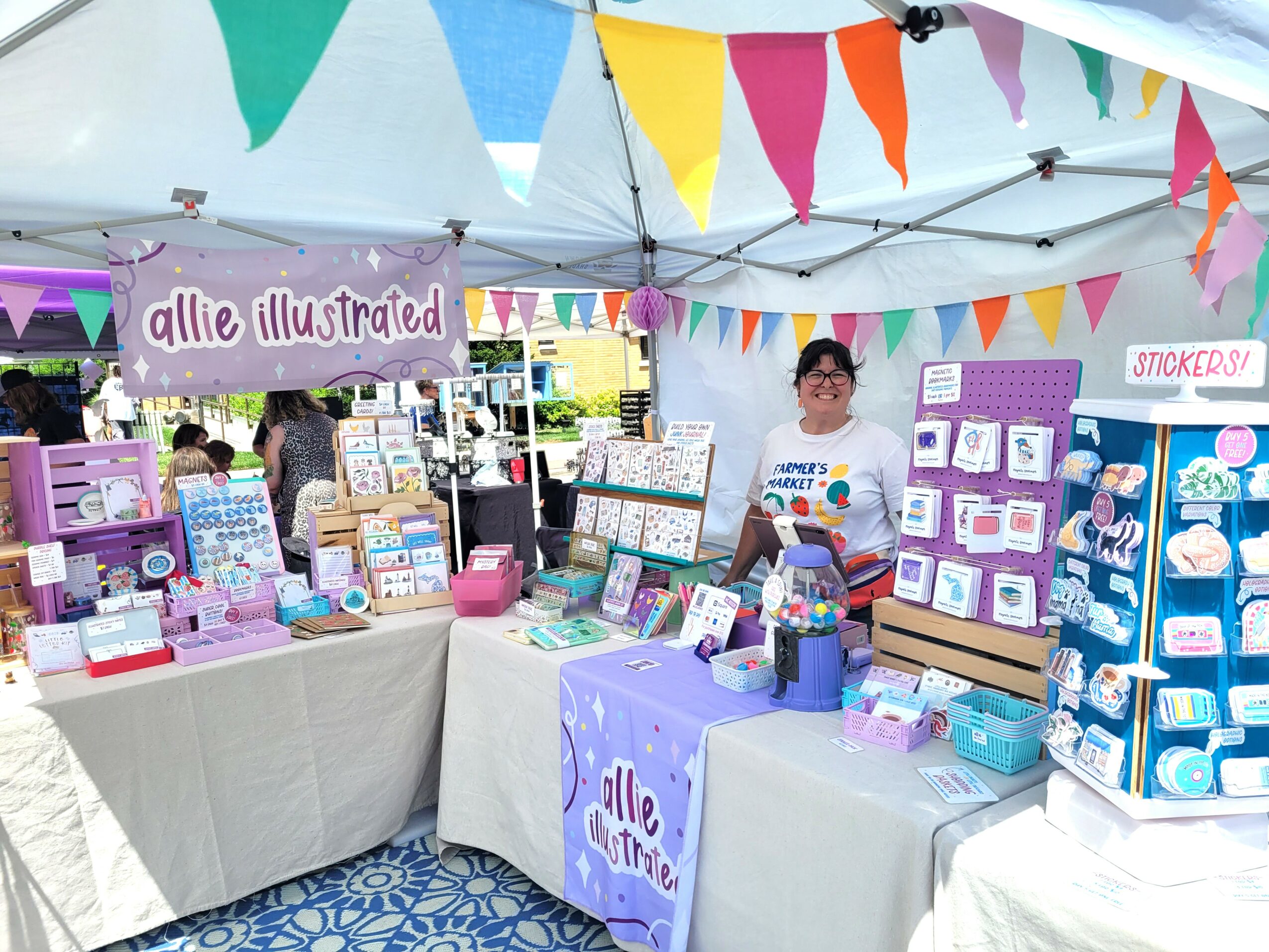 Vendor booth with colorful bunting, selling illustrated stationery and cards, with a person behind the table, under a tent.