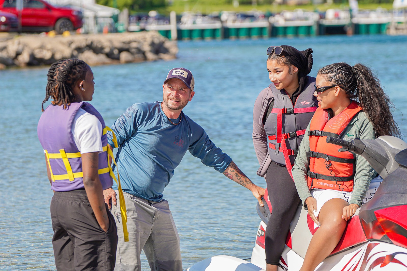 Four people in life jackets stand near water, engaging in conversation outdoors on a sunny day.