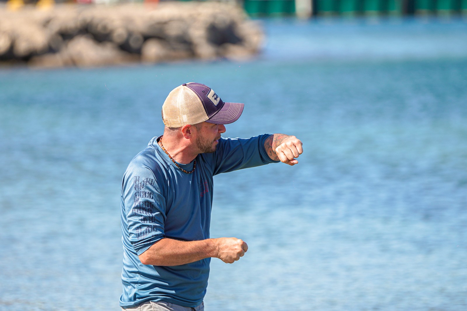 Man wearing a cap and blue shirt throws a punch near water, with rocks and a pier in background.
