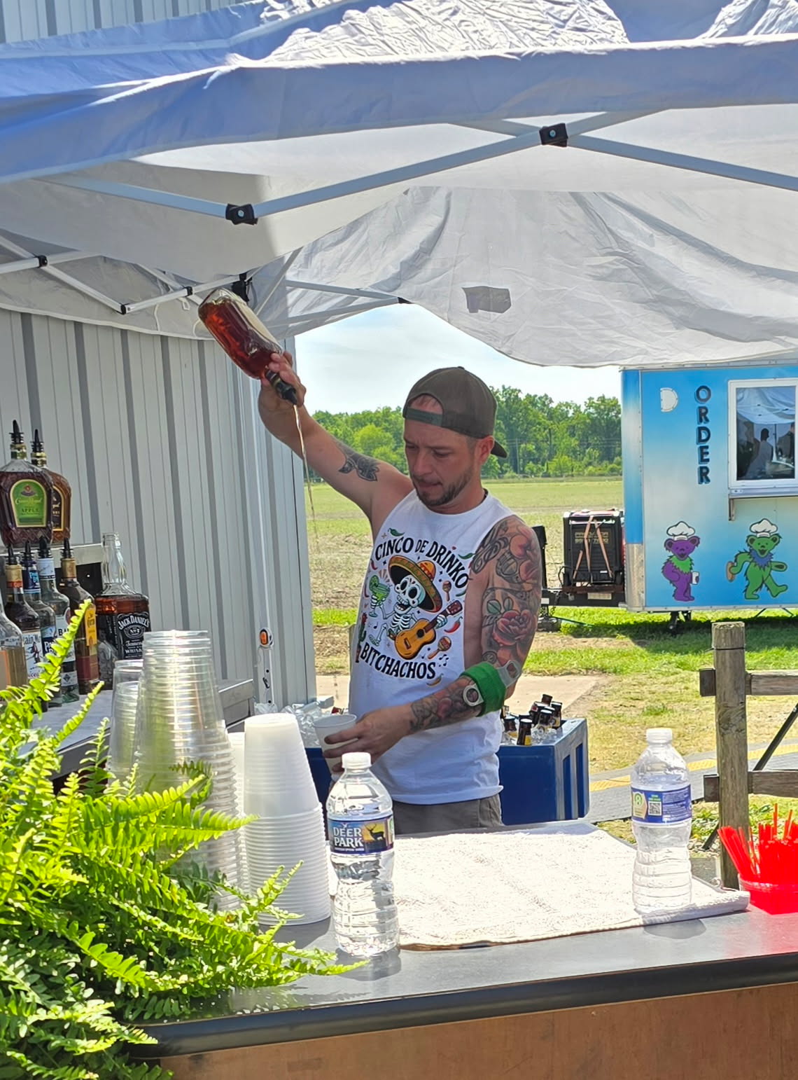 Man with tattoos and glasses pours a drink under a white canopy, with bottles, cups, and plants on the counter.
