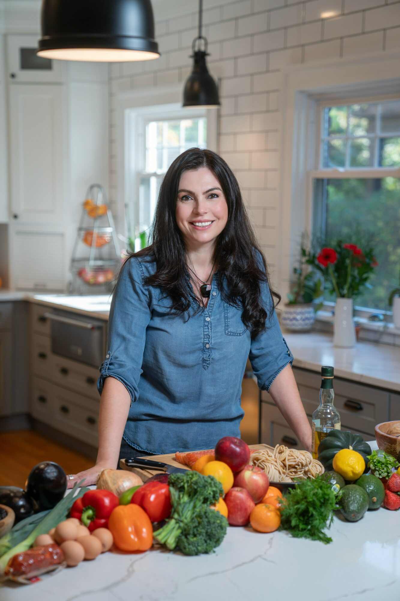 Woman with long dark hair in a denim shirt stands behind a kitchen counter with vegetables and fruits, smiling.