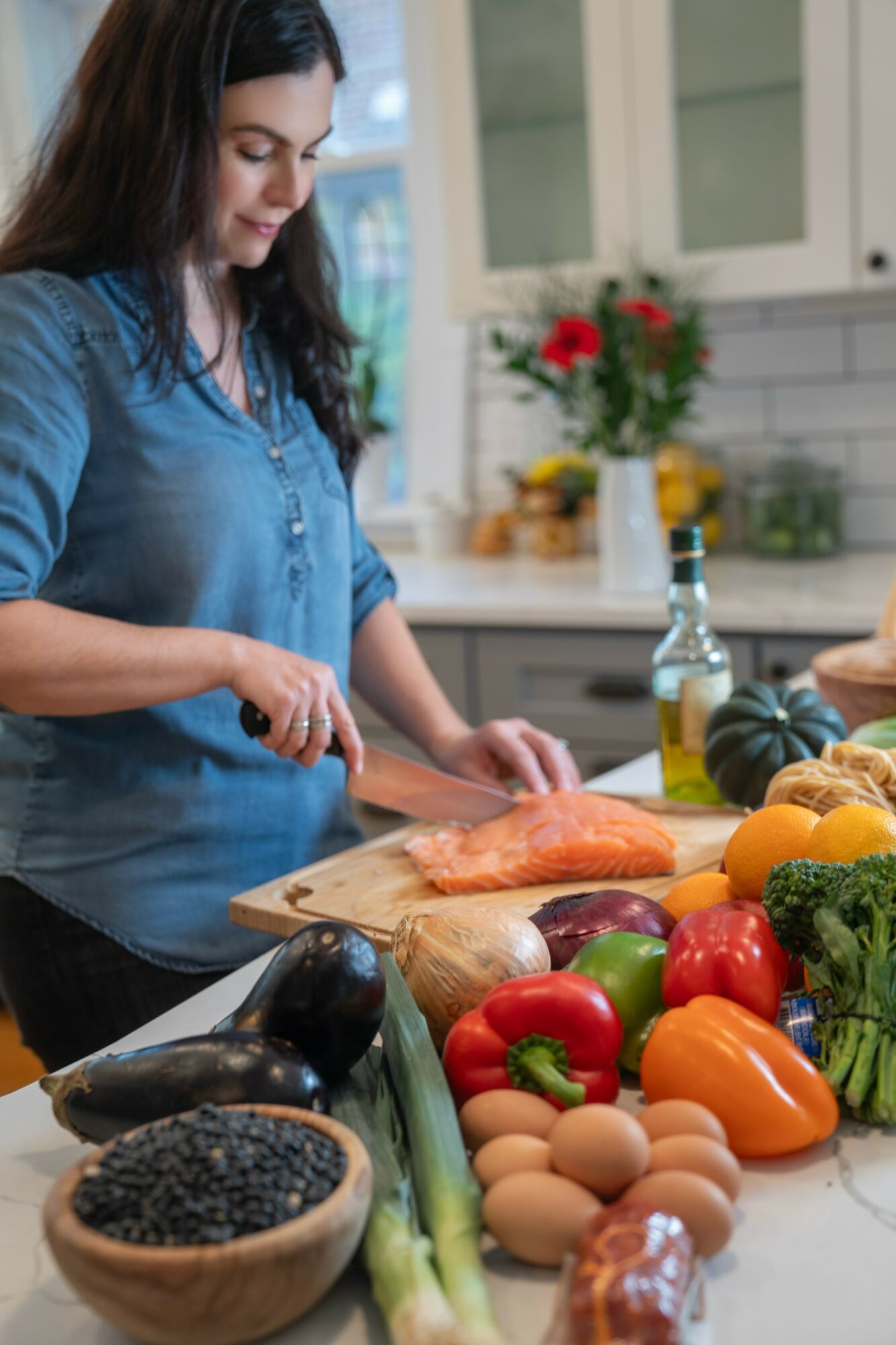 Woman preparing fish in a kitchen with vegetables, eggs, and cooking ingredients on the counter.