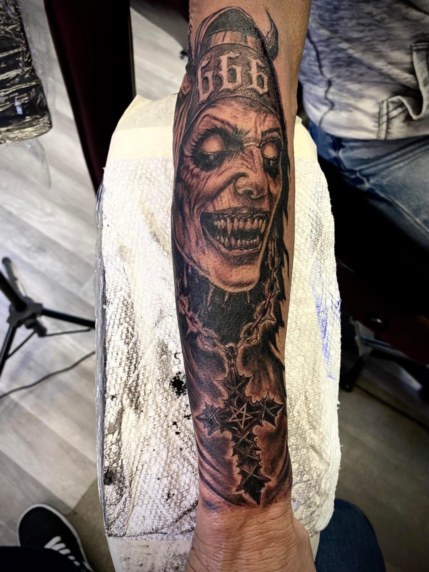 Tattoo of a grinning skull with a crown, surrounded by dark shading and decorative elements, on a person's forearm.