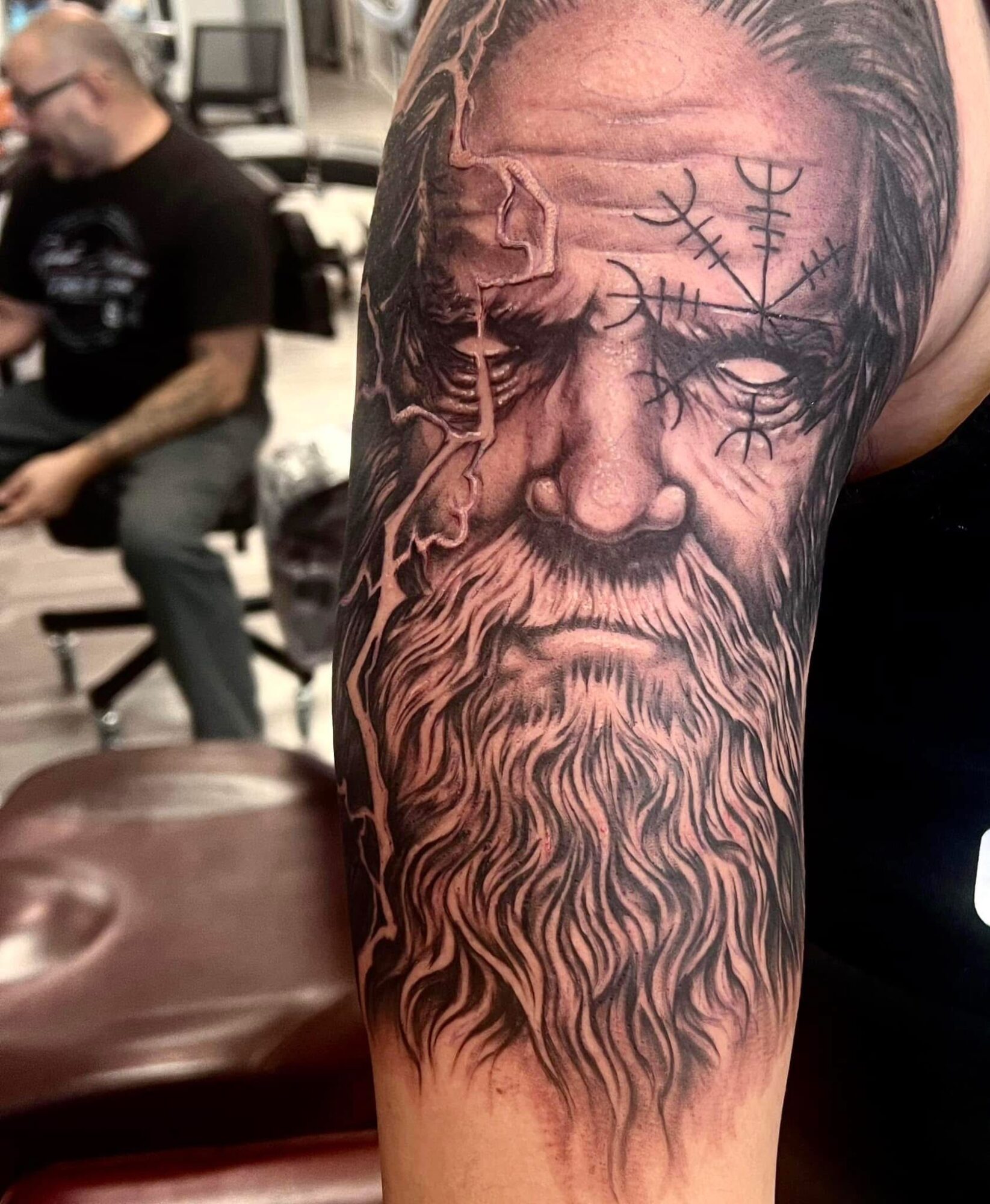 Close-up of a detailed black and gray tattoo of an elderly man's face with a long beard and symbols on his forehead.