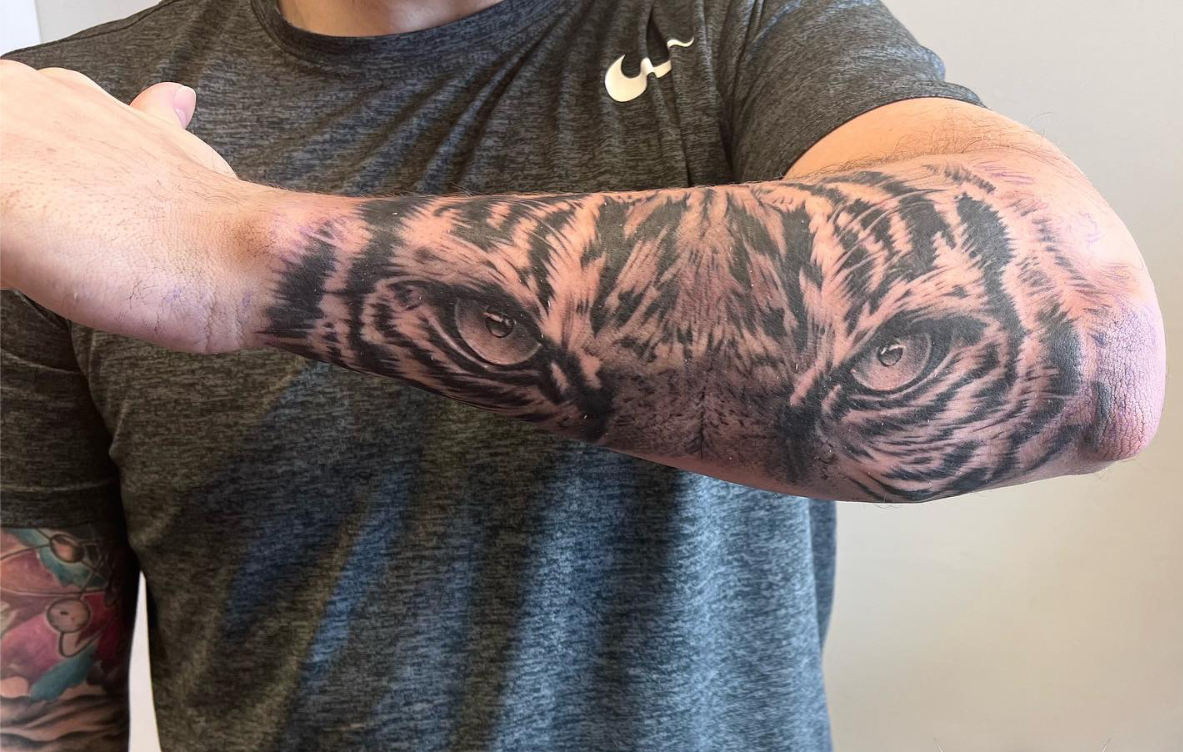 Person with a tiger face tattoo on their forearm, wearing a dark gray shirt, arm extended sideways.