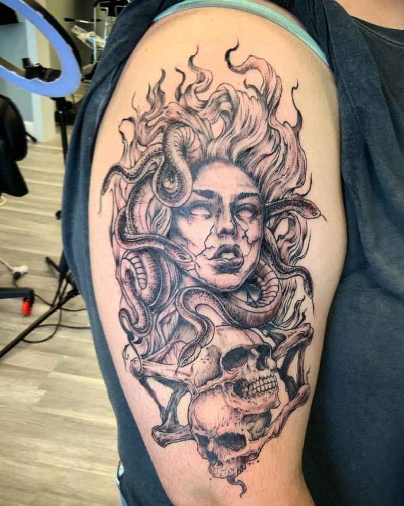 Black and gray tattoo of a woman's face with flowing hair, surrounded by snakes and a skull, on upper arm.