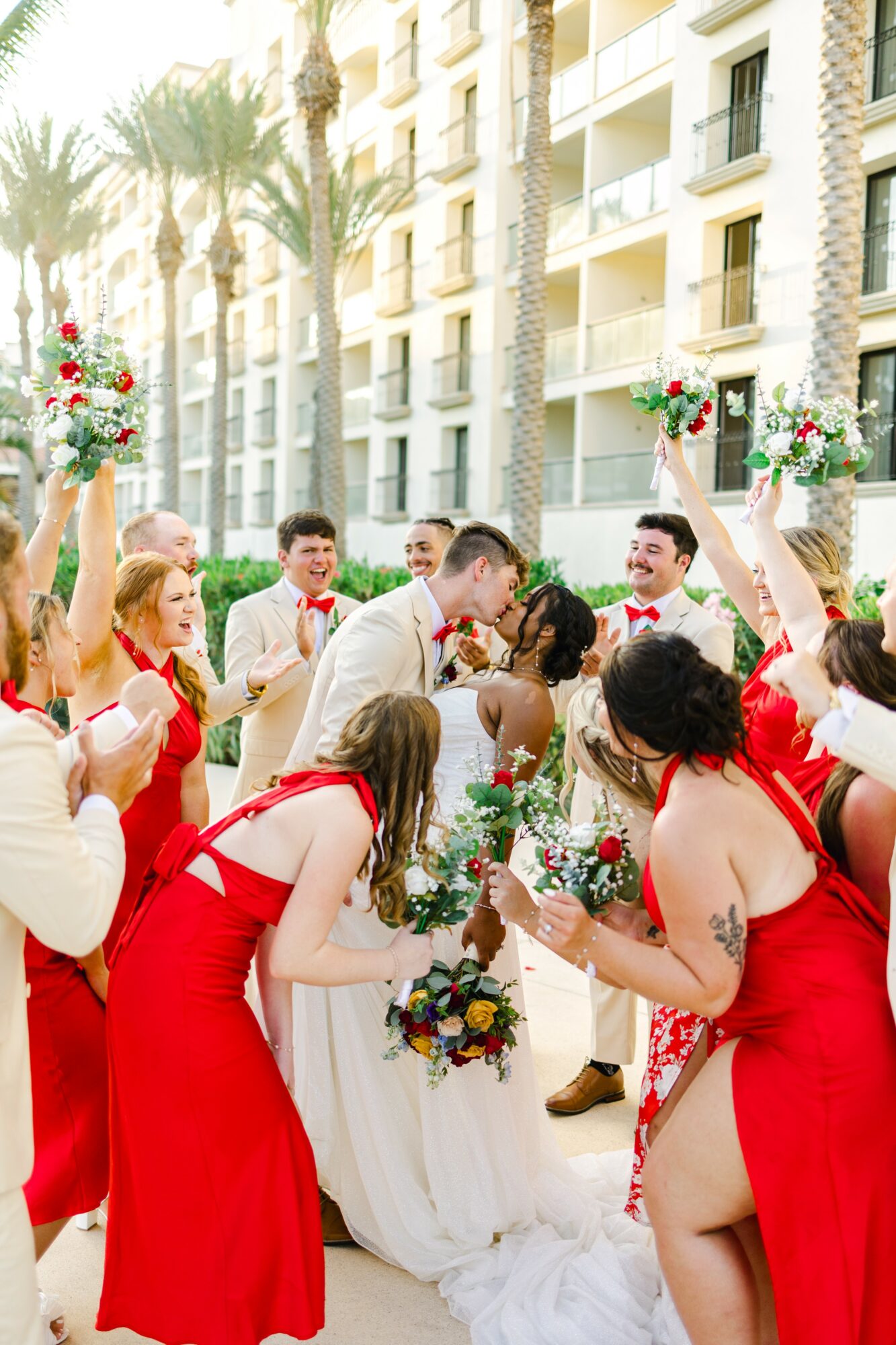 Group of people celebrating outdoors, some holding bouquets, in front of a building with palm trees, wearing red and white clothing.
