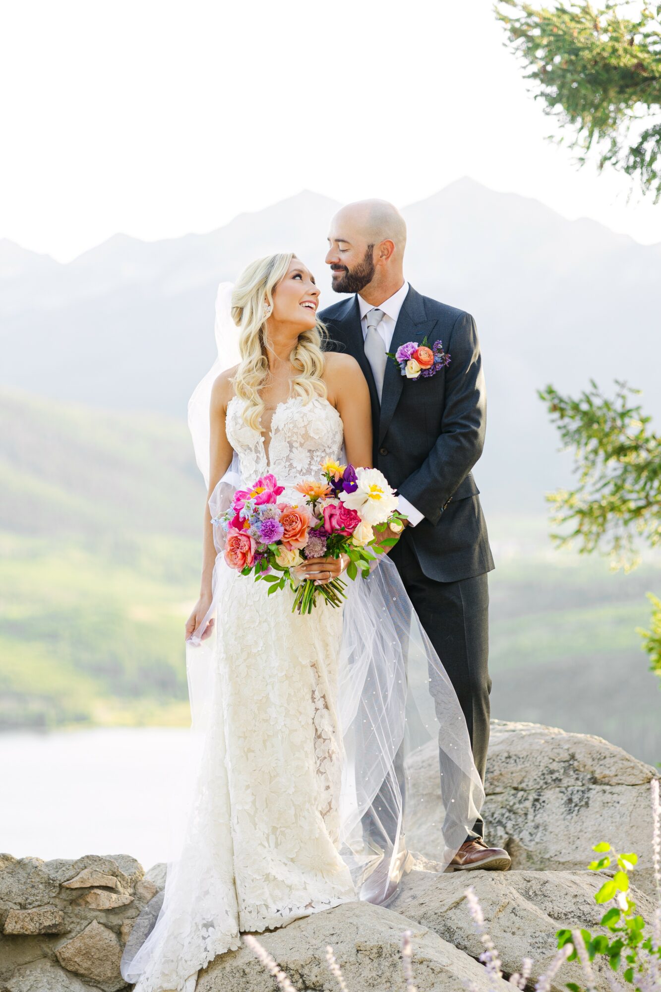 Bride and groom stand outdoors with mountains in background, holding a colorful bouquet, smiling at each other.