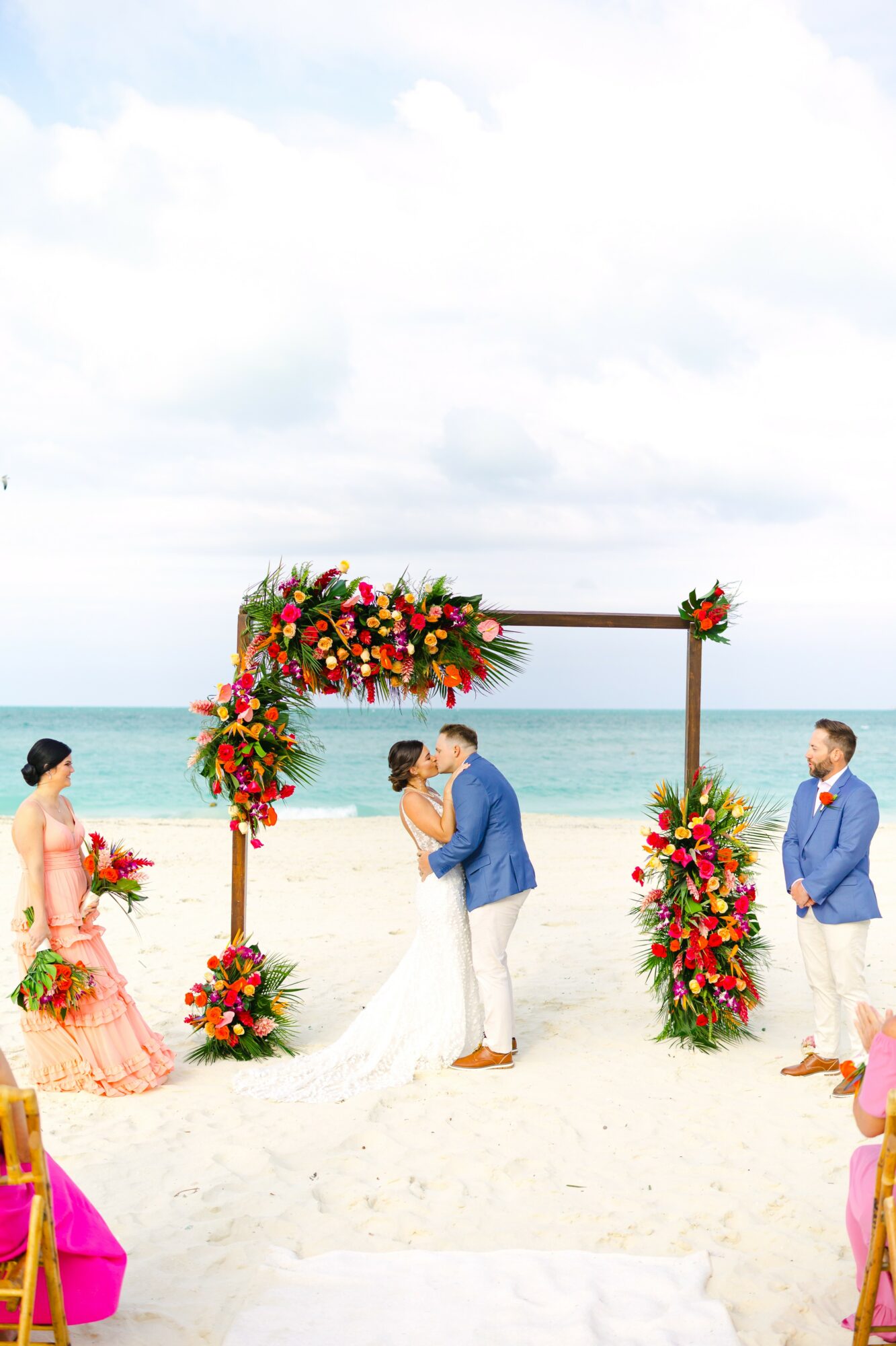 Couple exchanging vows under a decorated arch on a beach, with two people on each side, ocean in background.