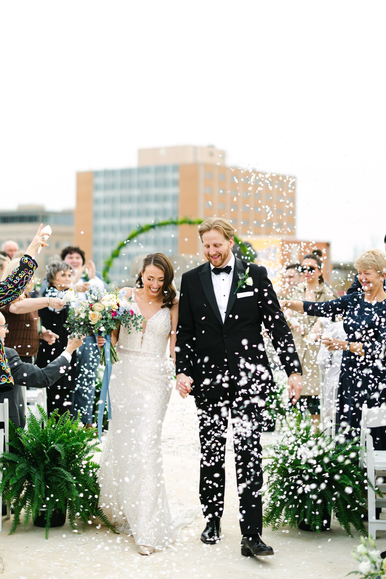 Bride and groom walking outdoors, surrounded by celebrating guests throwing confetti, with buildings in the background.