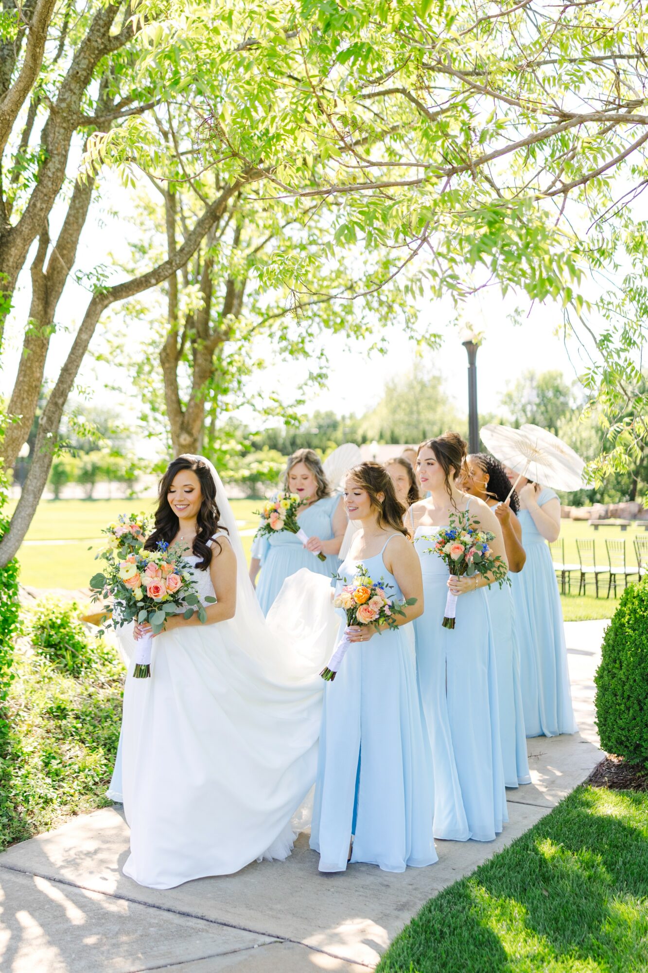 Bride in white dress with four bridesmaids in light blue dresses holding bouquets outdoors under a tree.