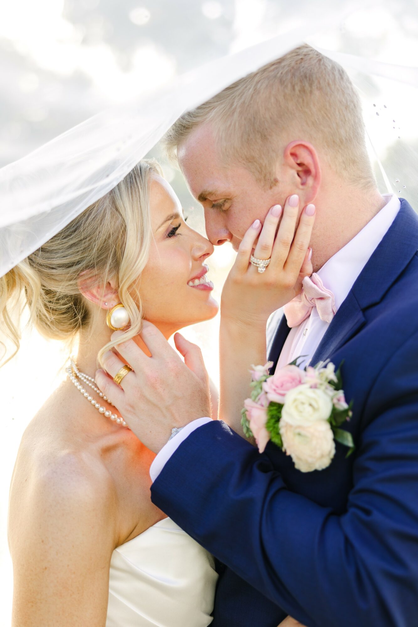 Bride and groom close together, smiling, with bride touching groom's face, wedding attire, veil, and bouquet visible.