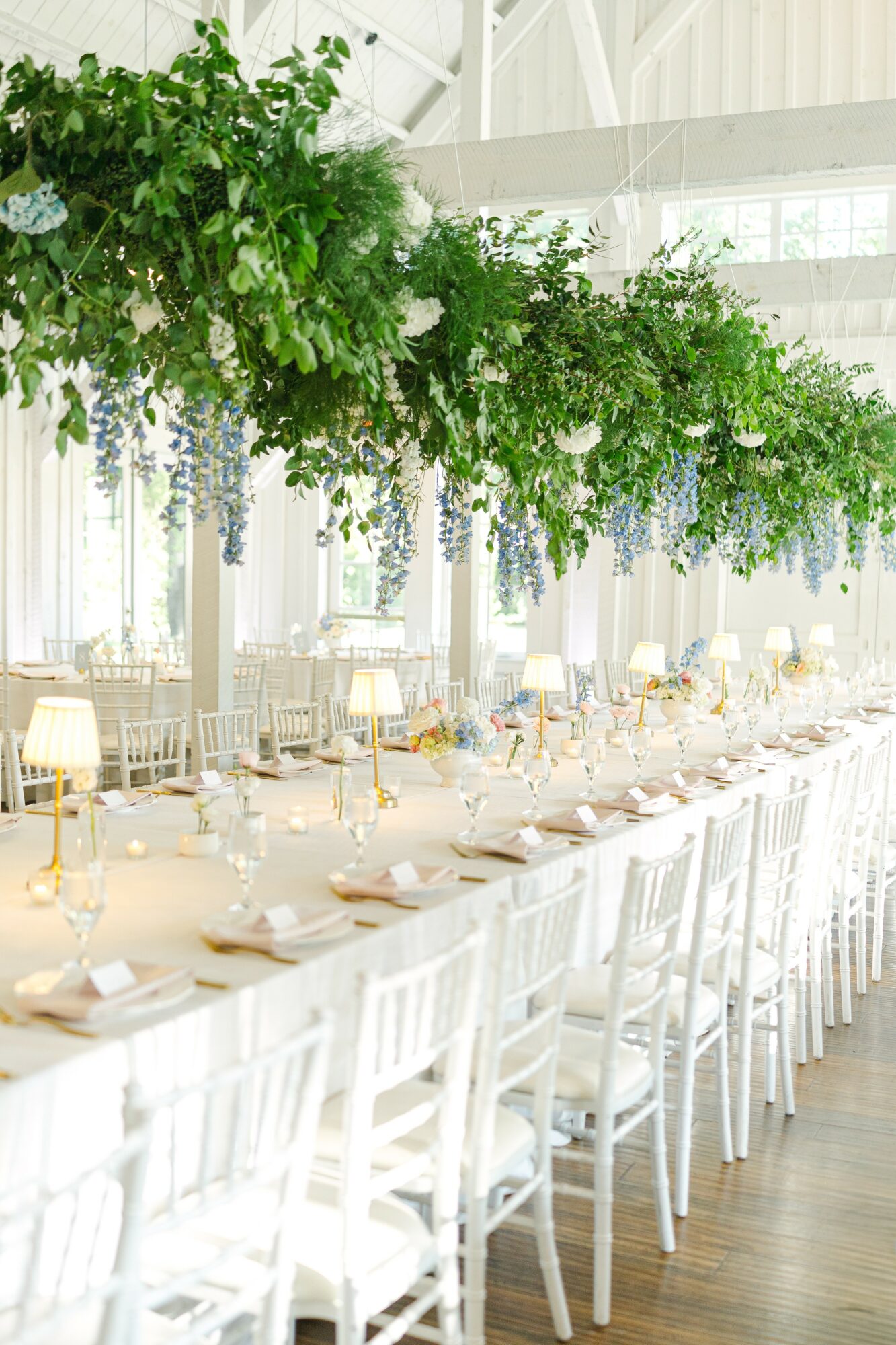 Long banquet table decorated with white chairs, floral arrangements, and table settings in a bright, airy venue.