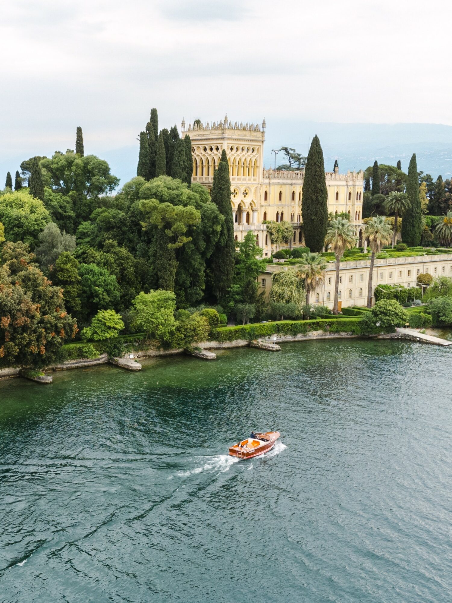A boat moves across a body of water with a large historic building and lush trees in the background.